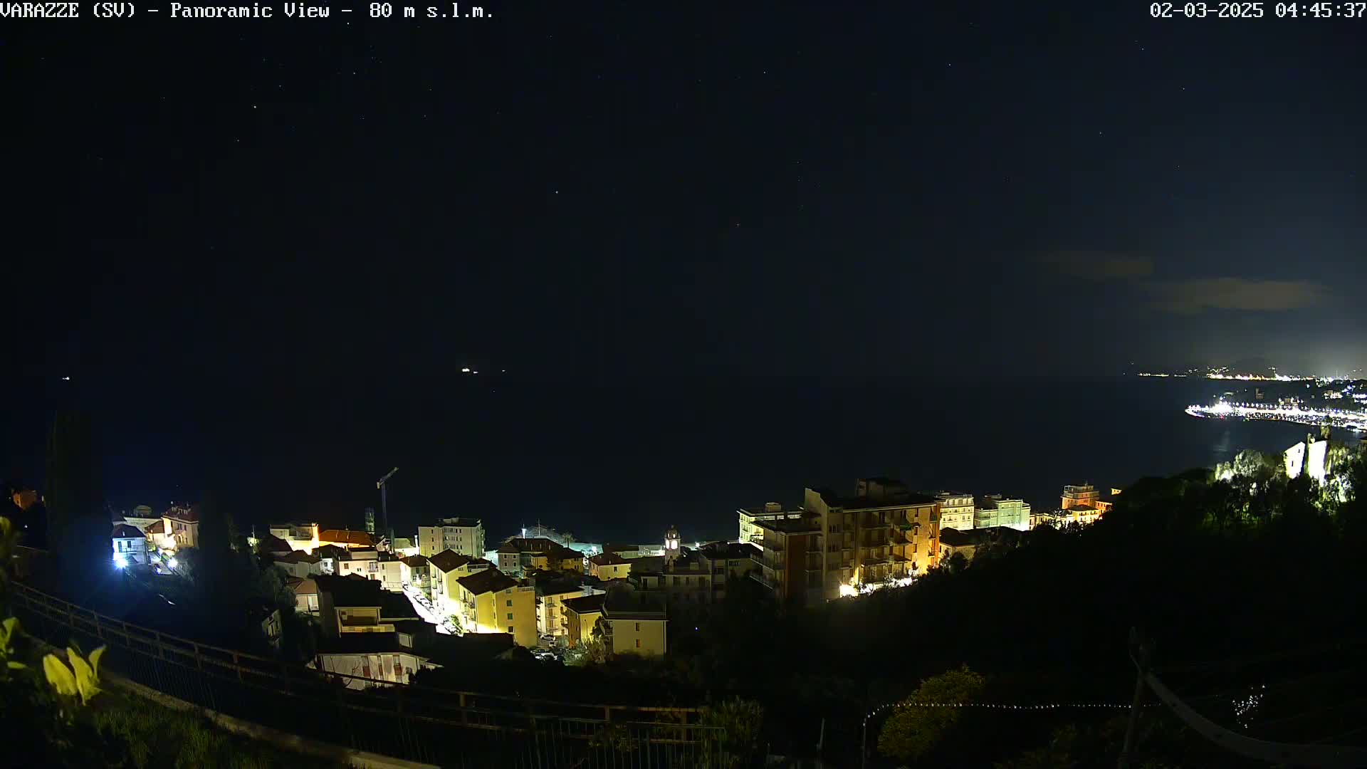 A nighttime panoramic view shows a coastal town with its lights illuminating the buildings, situated on a hill overlooking a dark sea under a clear, starry sky.
