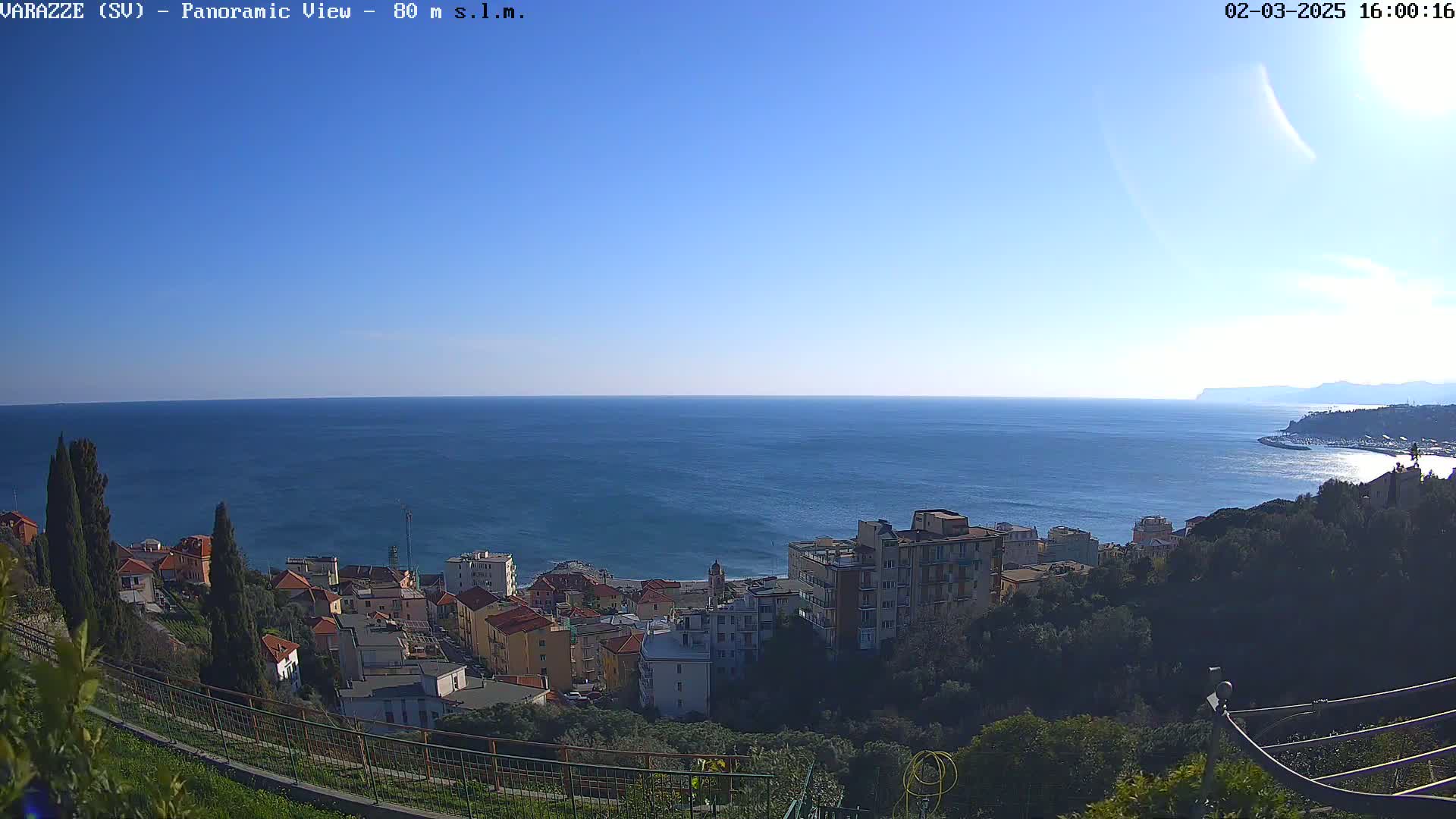 A coastal town sits on a hillside overlooking a calm, blue sea under a clear blue sky.
