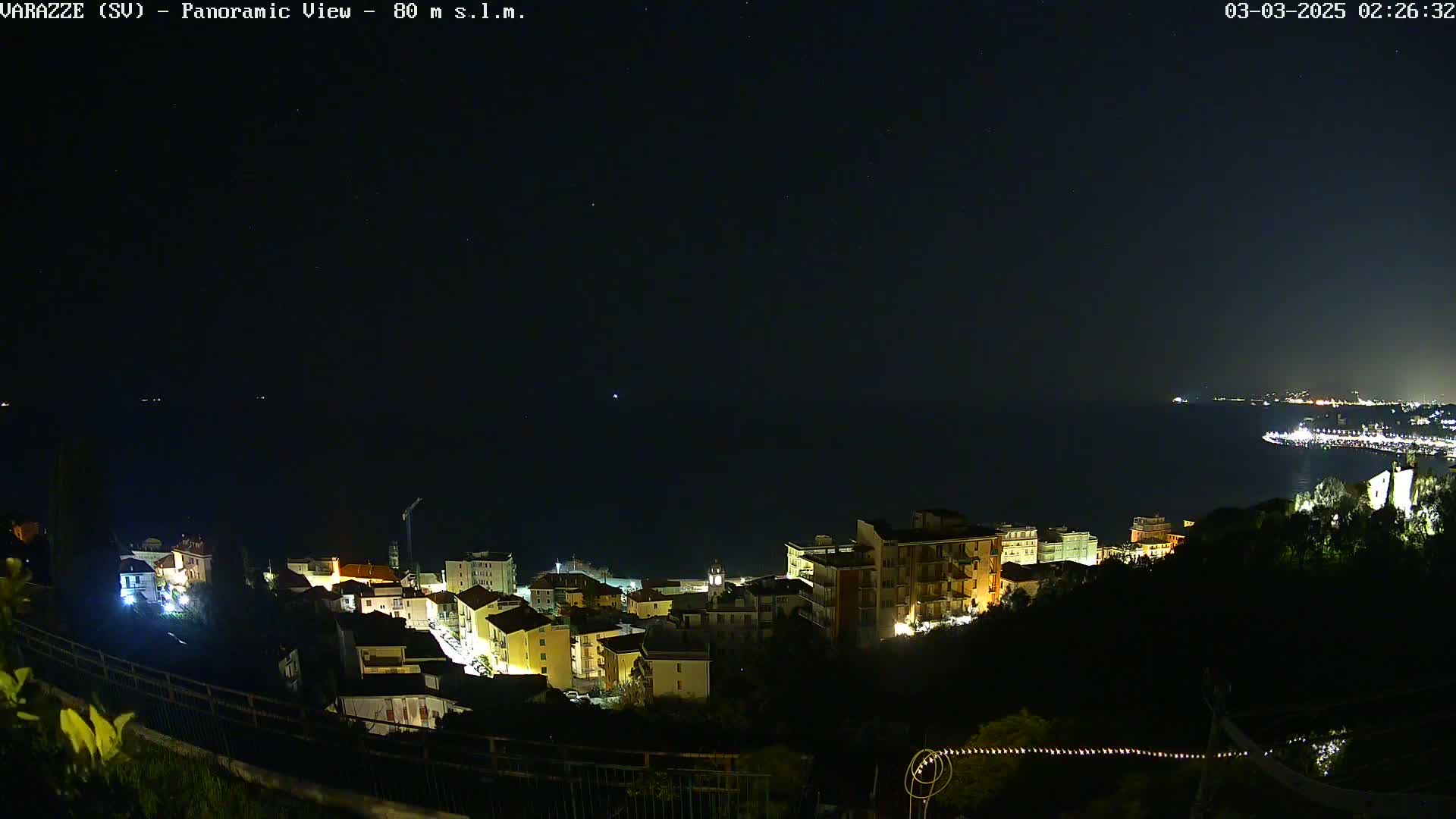 A nighttime, panoramic view of a coastal town with buildings illuminated, overlooking a dark body of water under a clear, starry sky.