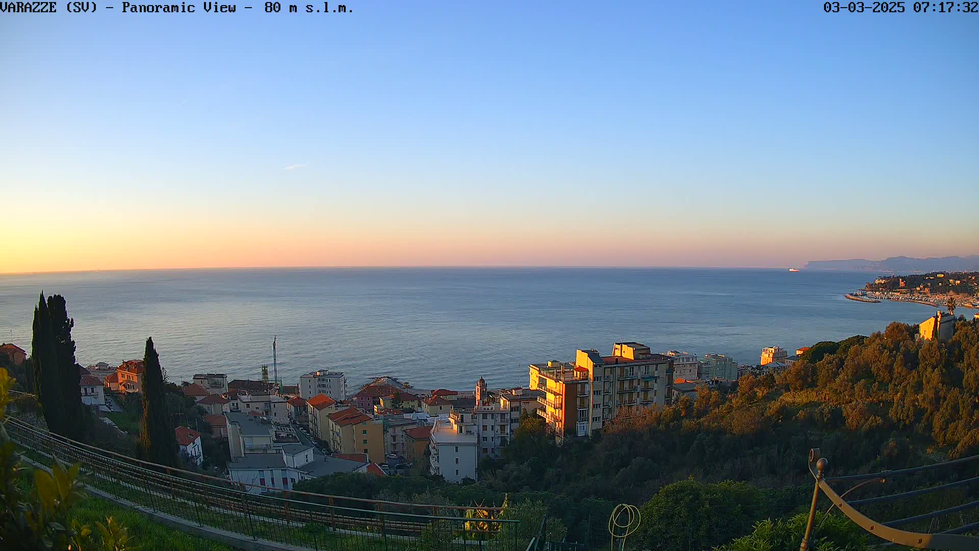 A coastal town nestled on a hillside overlooks a calm sea during a sunset under a clear sky.