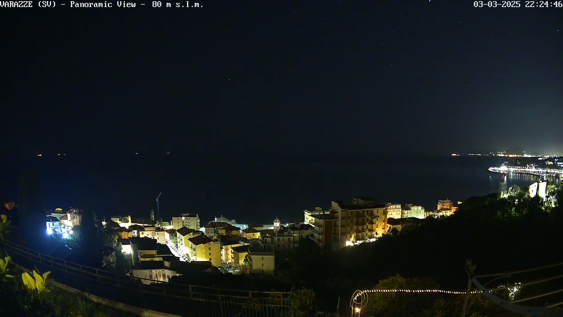 A nighttime panoramic view shows a coastal town illuminated, with a dark sea and starry sky in the background.