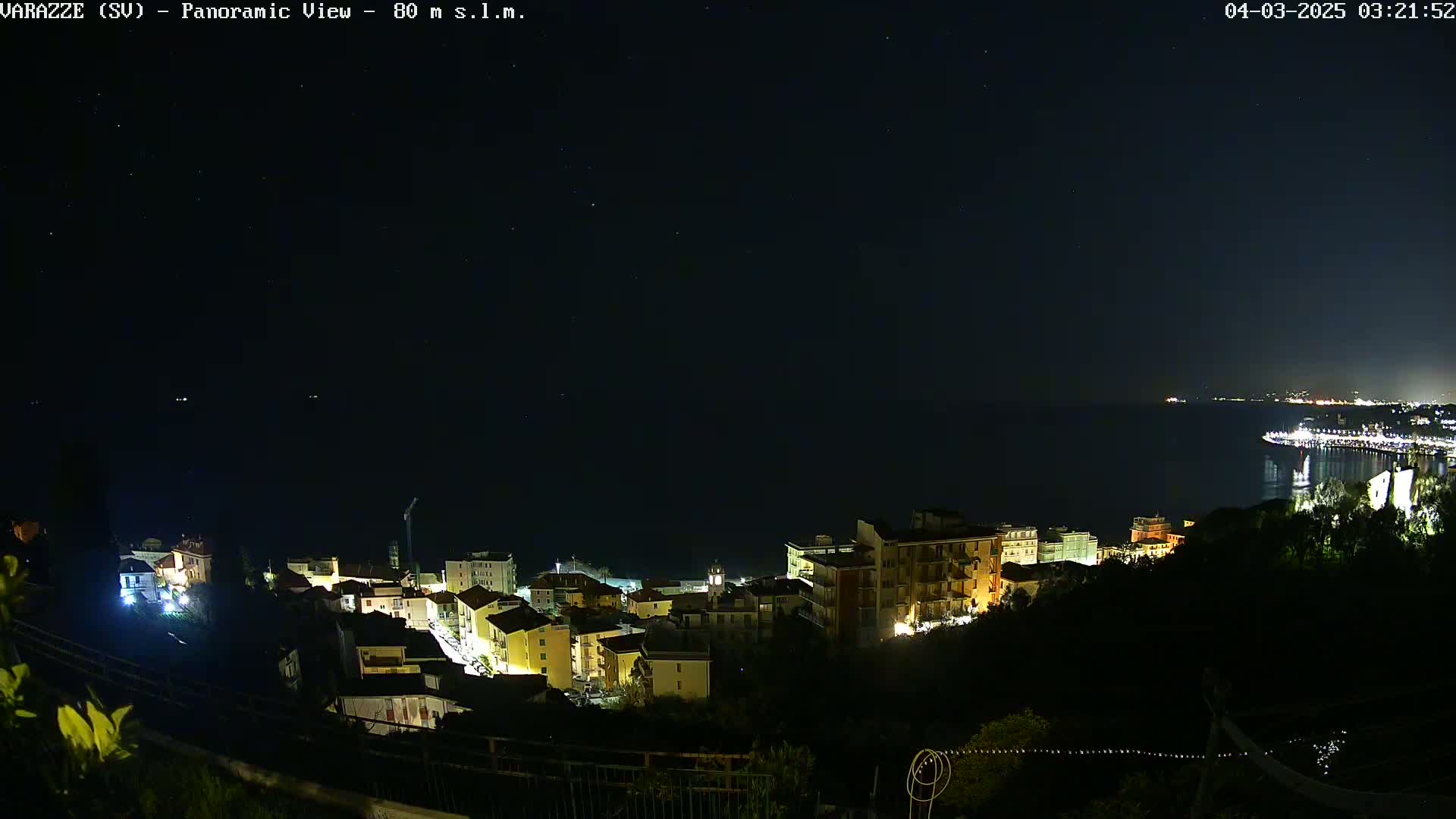 A nighttime, panoramic view of a coastal town illuminated with lights, overlooking a dark body of water under a clear, starry sky.