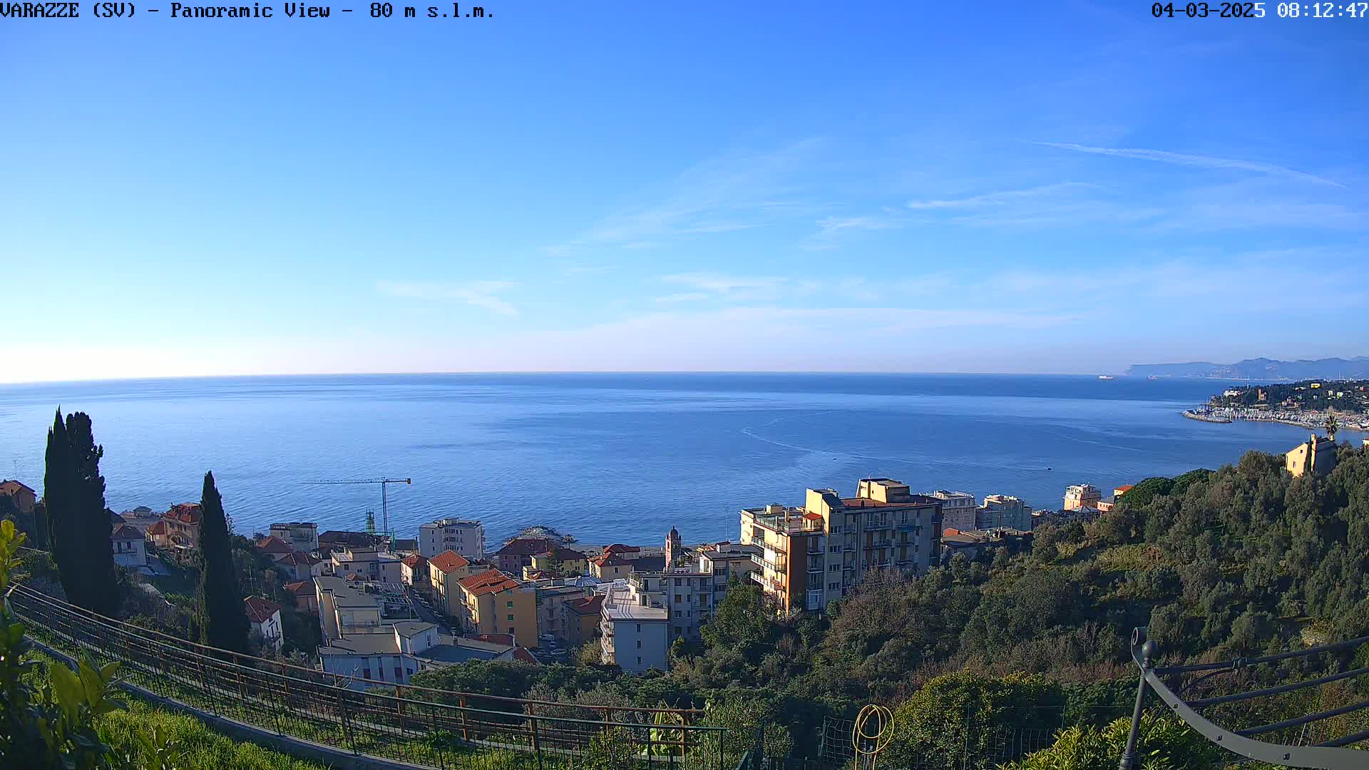 A panoramic view of a coastal town nestled on a hillside overlooking a calm, blue sea under a clear blue sky.