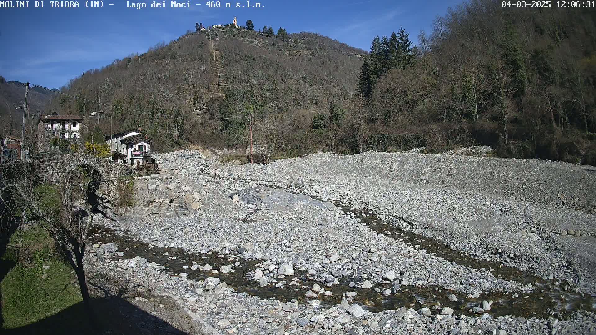 A rocky, mostly dry riverbed with a small stream flows through it, nestled between a stone bridge and a hillside covered in leafless trees under a clear, sunny sky.