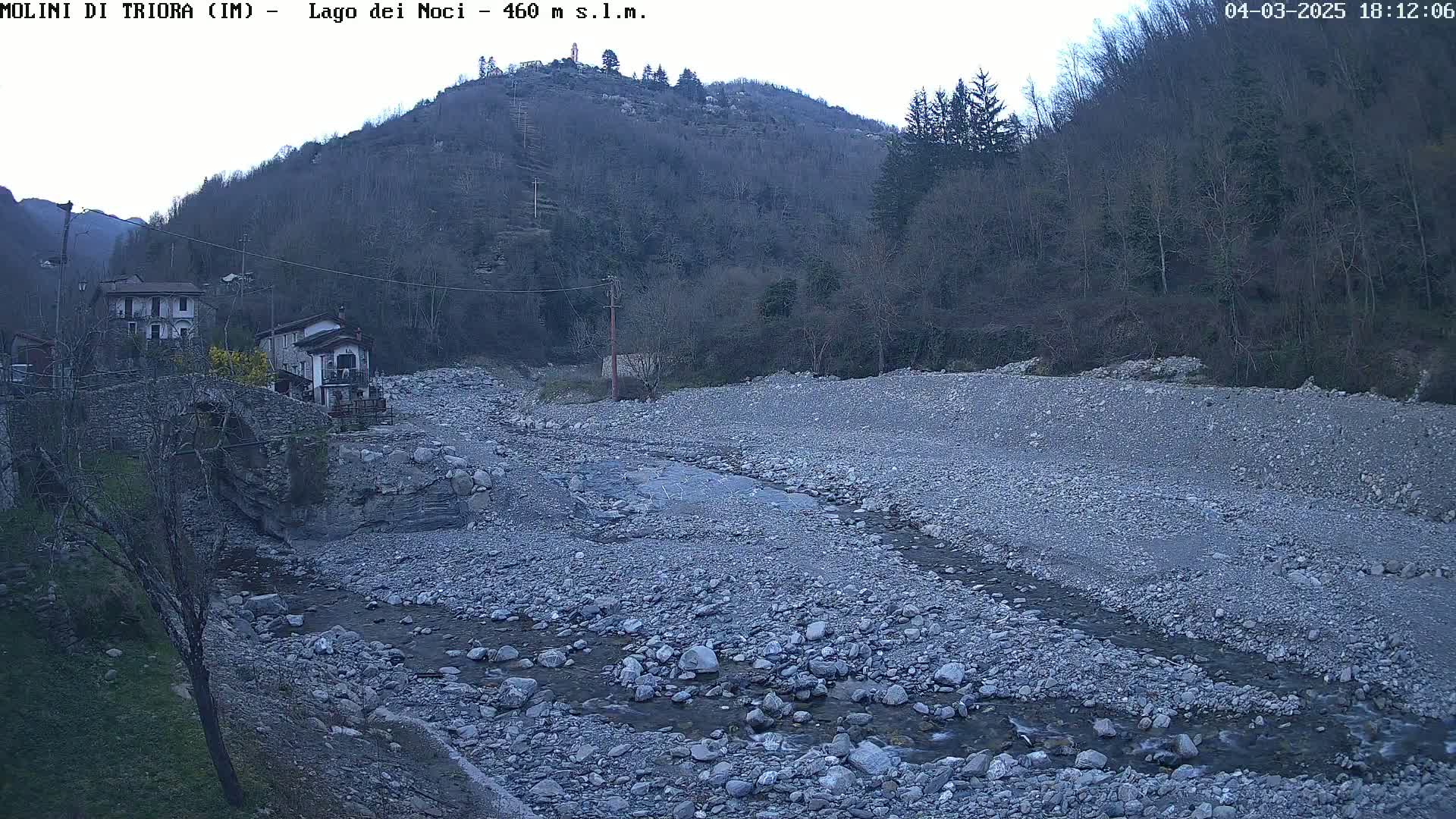 A rocky, mostly dry riverbed winds through a valley under an overcast sky, past a stone bridge and a couple of houses nestled at the base of a sparsely wooded hill.