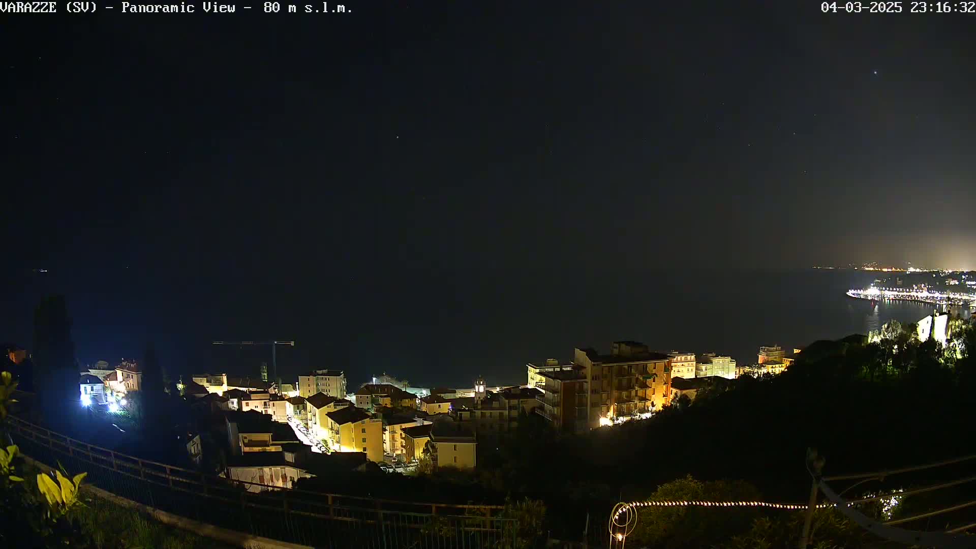 A nighttime panoramic view of a coastal town illuminated by lights, with a dark sky showing some stars and a calm sea in the background.