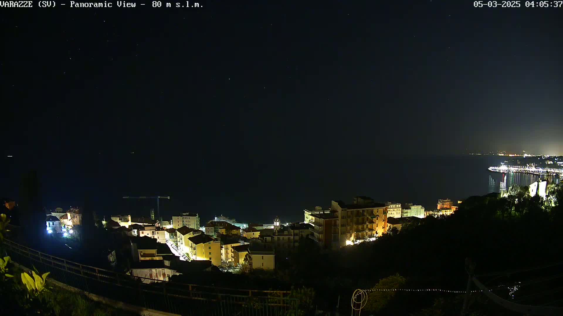A nighttime panoramic view shows a coastal town illuminated with lights, overlooking a dark body of water under a clear, starry sky.
