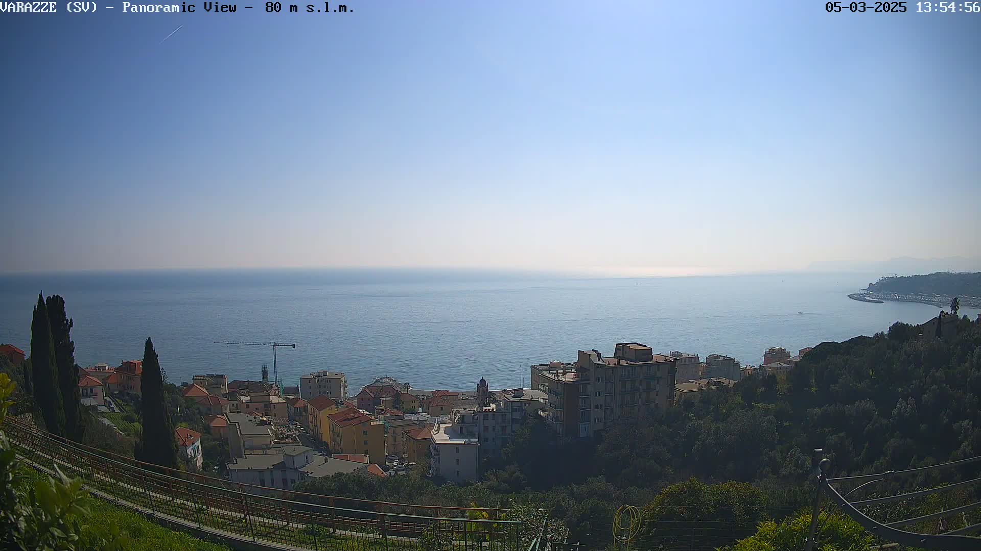 A coastal town nestled on a hillside overlooking a calm, blue sea under a clear, sunny sky.