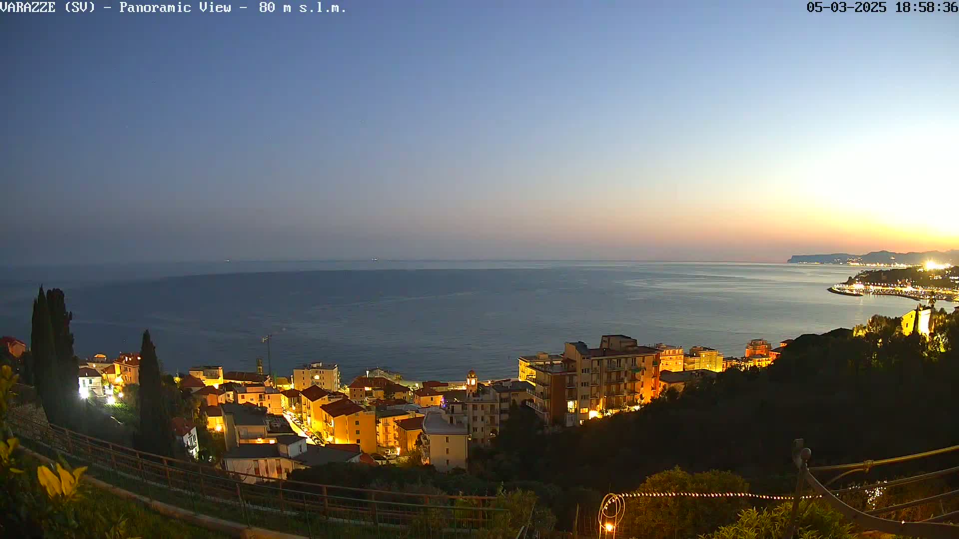 A coastal town with illuminated buildings at dusk overlooks a calm, dark-blue sea under a twilight sky.