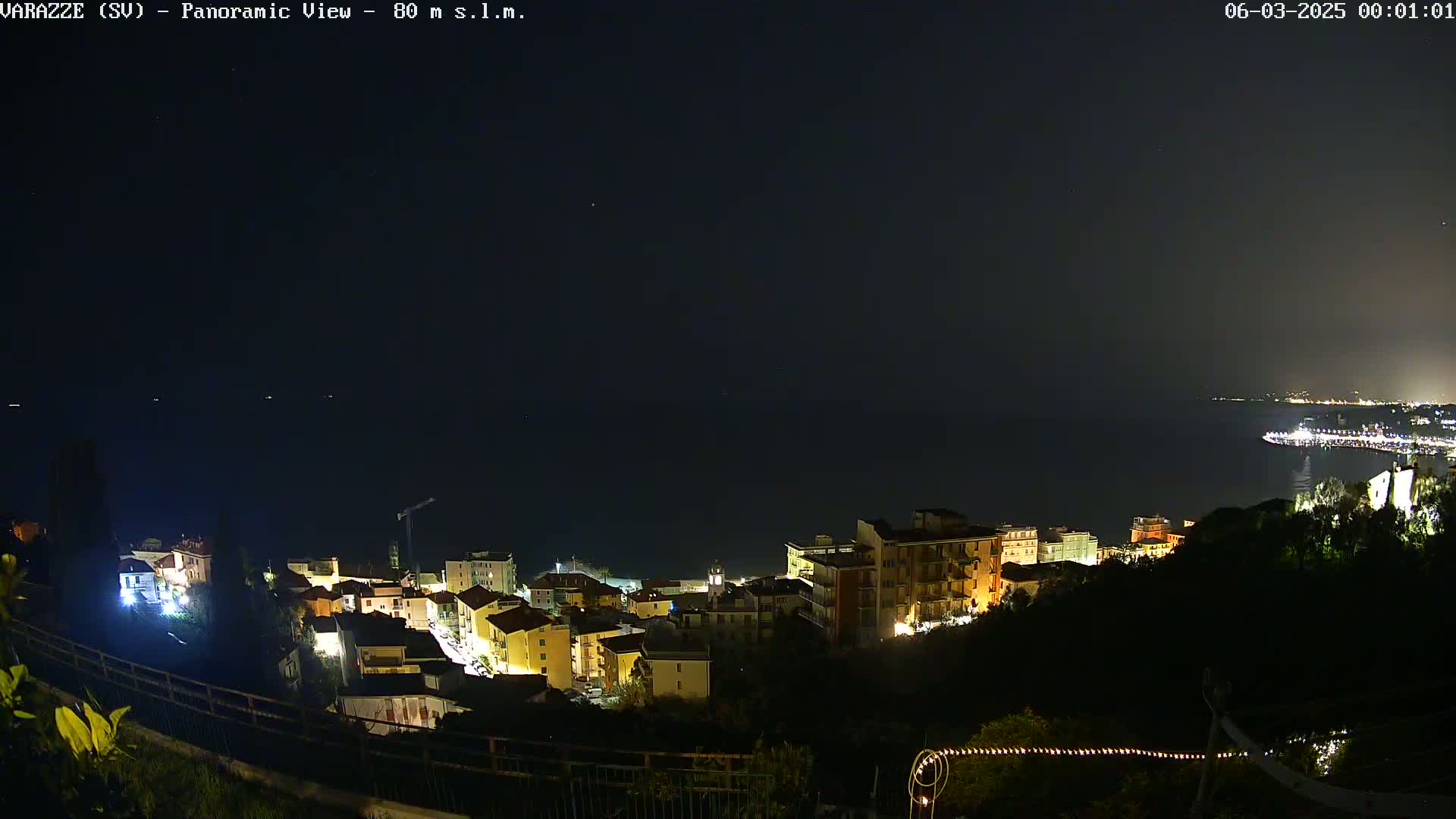 A nighttime, panoramic view of a coastal town illuminated with lights, overlooking a dark body of water under a clear, starry sky.