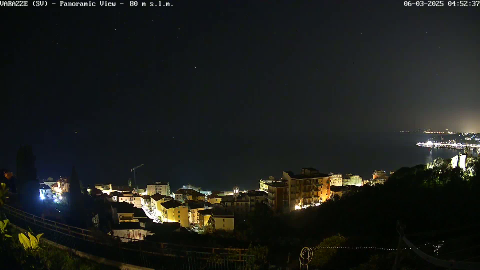 A nighttime, panoramic view of a coastal town with its lights illuminated, overlooking a dark body of water under a clear, starry sky.