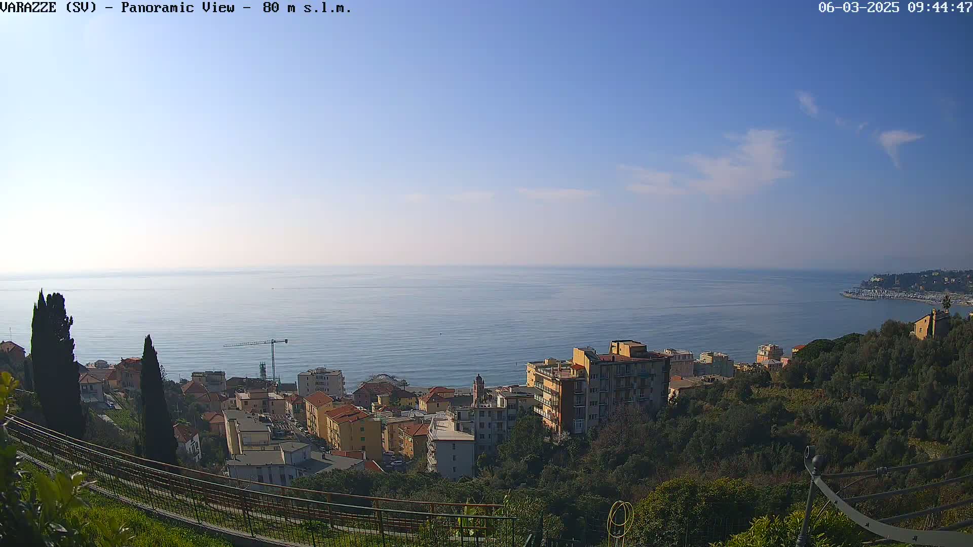 A panoramic view of a coastal town under a mostly clear, sunny sky, showing a calm sea and buildings nestled amongst green hills.