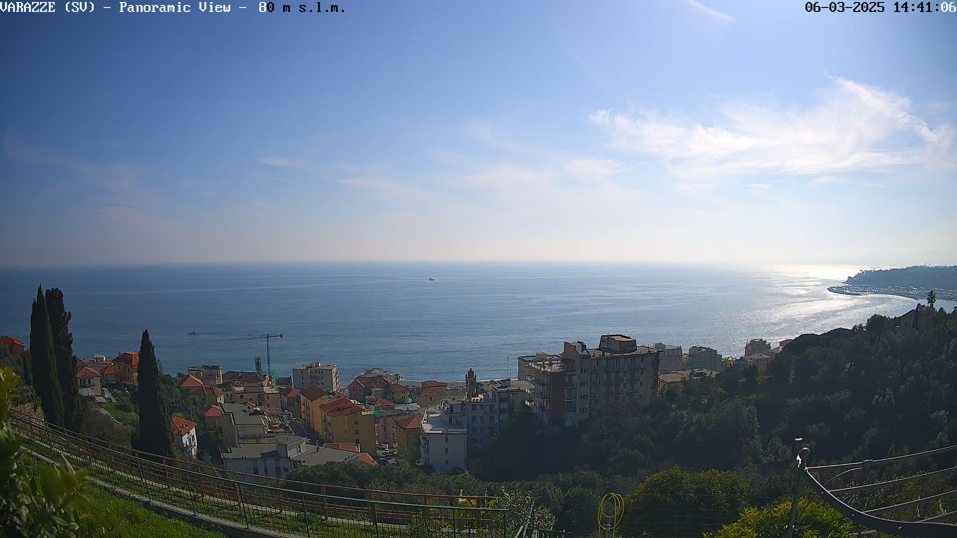 A panoramic view shows a coastal town nestled on a hillside overlooking a calm sea under a mostly clear sky.