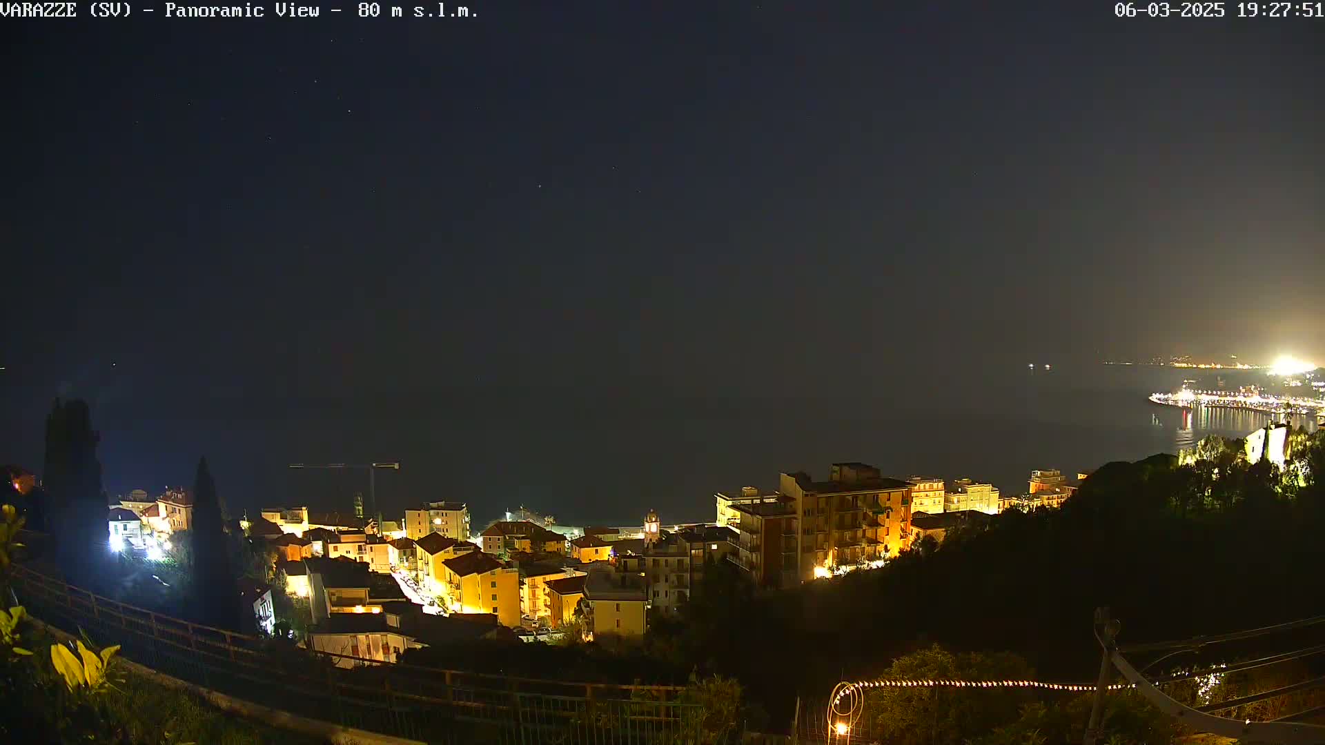 A nighttime, panoramic view of a coastal town with its buildings illuminated, overlooking a dark sea under a clear, starry sky.