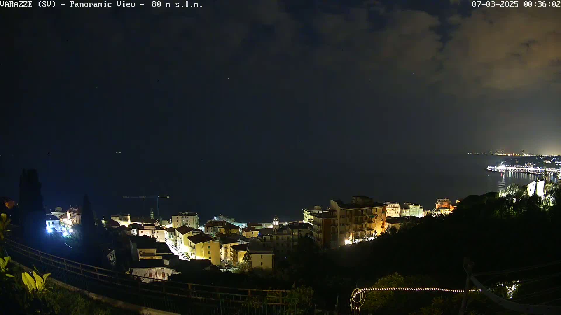A nighttime panoramic view of a coastal town with buildings lit up, under a partly cloudy sky, overlooking a dark body of water.