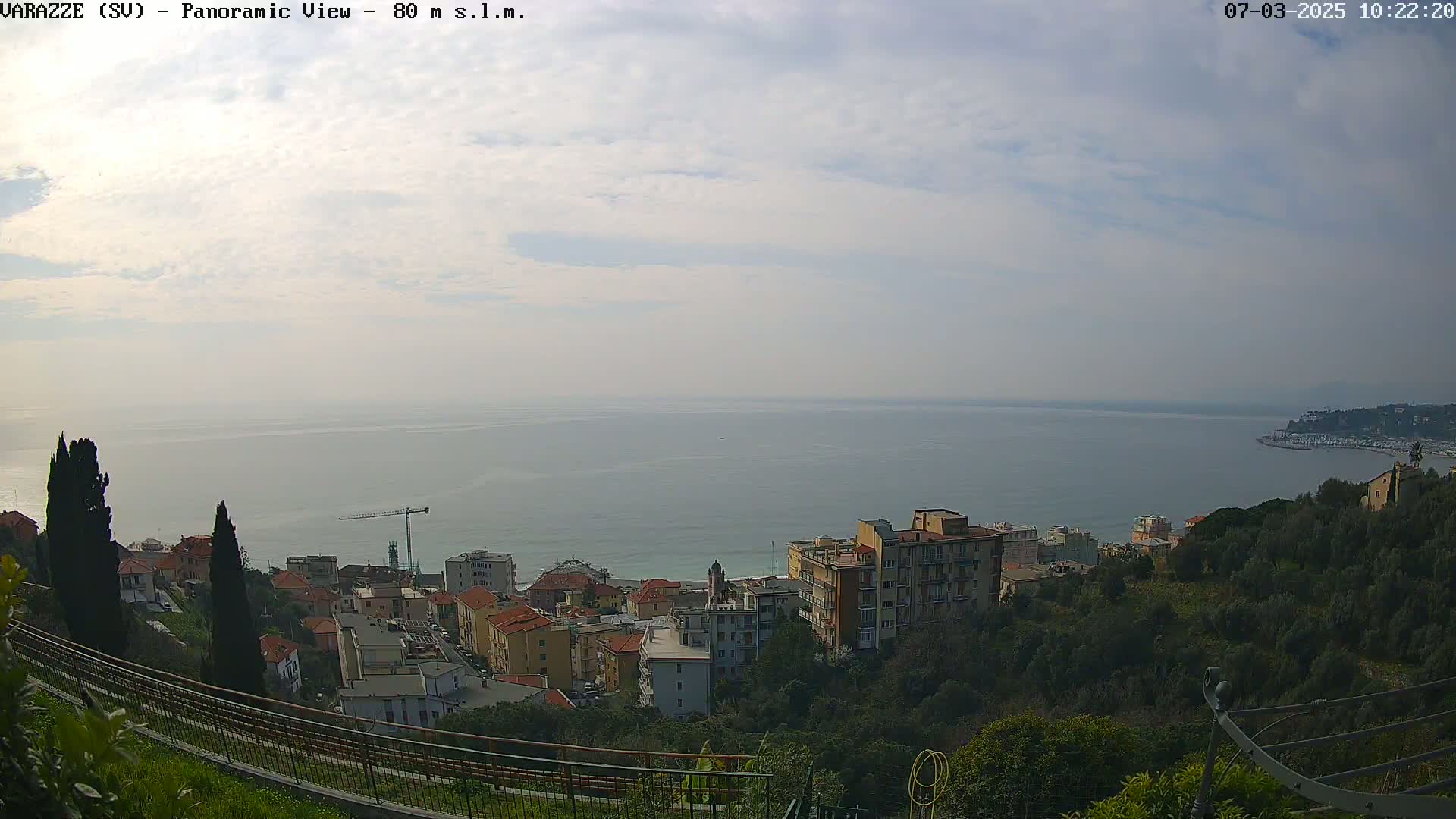 A coastal town nestled on a hillside overlooks a calm sea under a mostly cloudy sky.