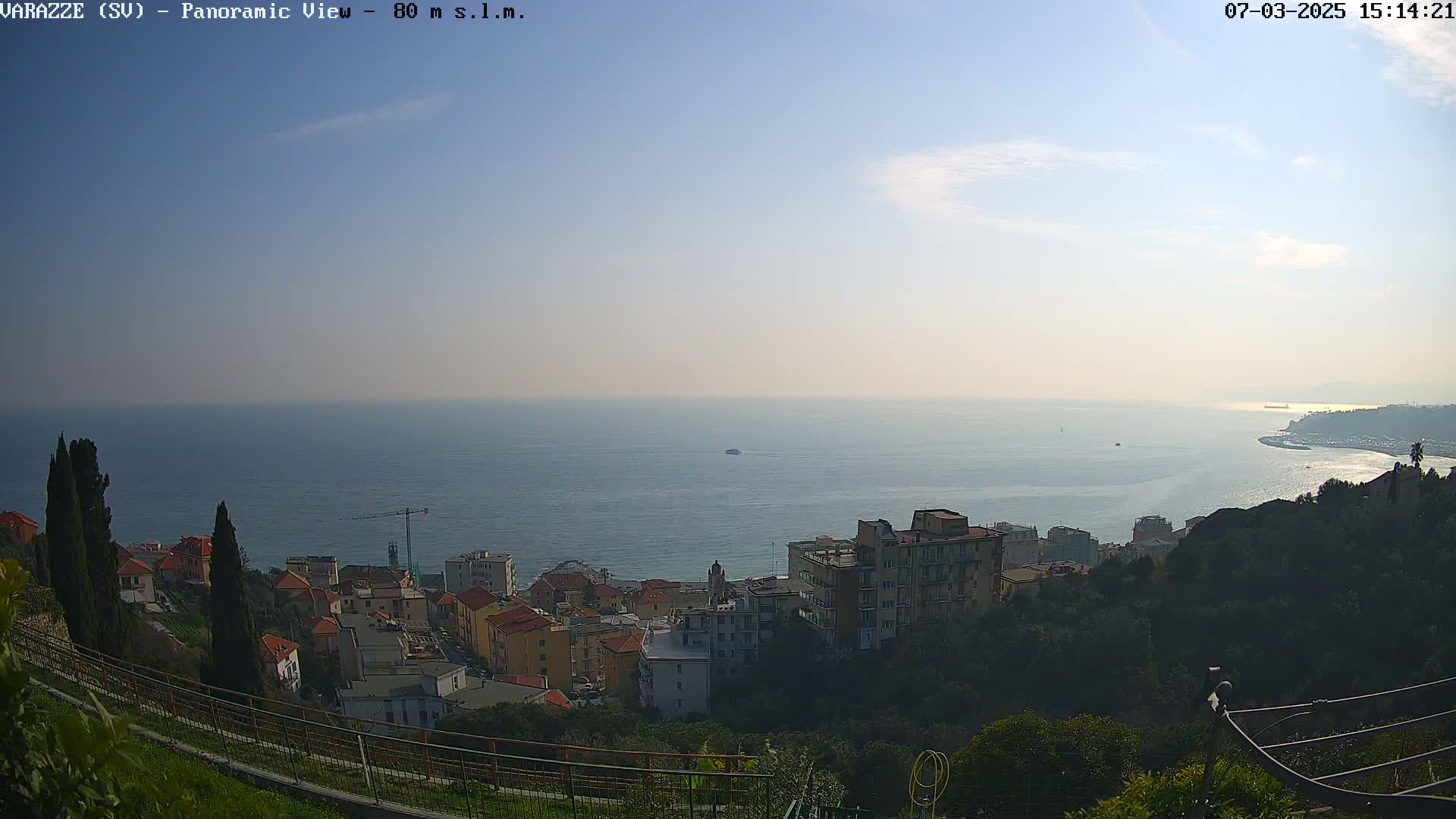 A panoramic view shows a coastal town nestled on a hillside overlooking a calm sea under a mostly clear sky.