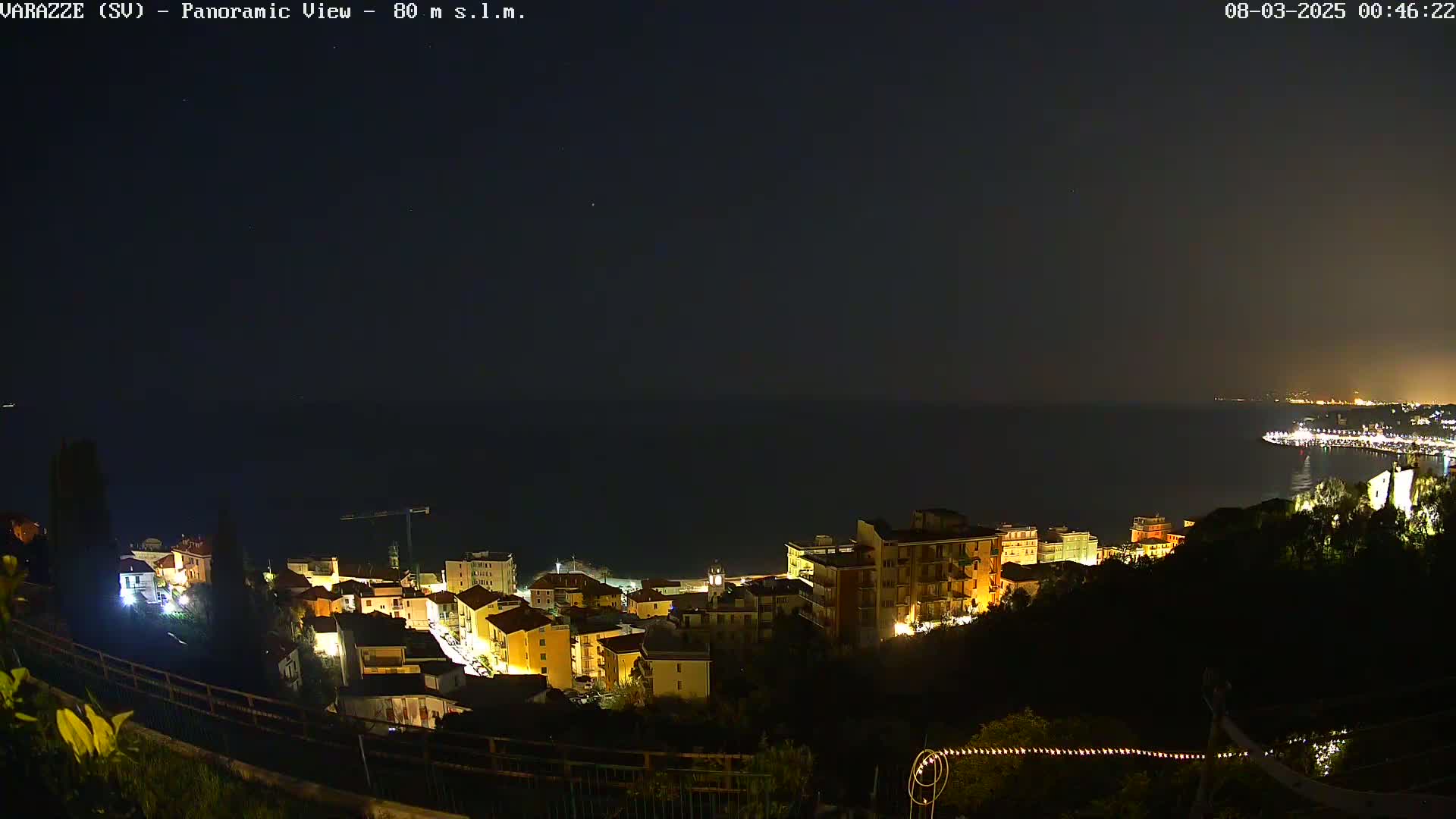 A nighttime panoramic view shows a coastal town illuminated, nestled on a hillside overlooking a dark sea under a clear sky.