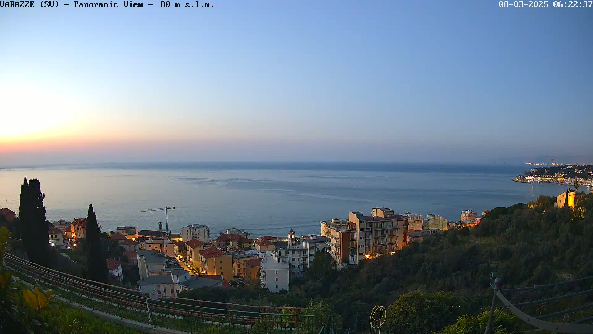 A coastal town at twilight is situated on a hillside overlooking a calm sea under a clear, dusky sky.