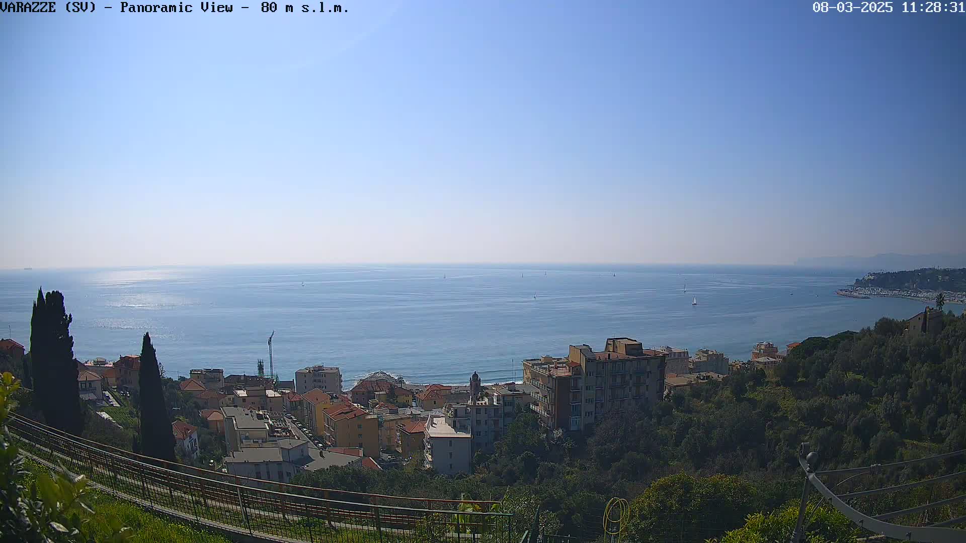 A panoramic view shows a clear, sunny day over a calm ocean with several sailboats, overlooking a hillside town with low buildings and green trees.