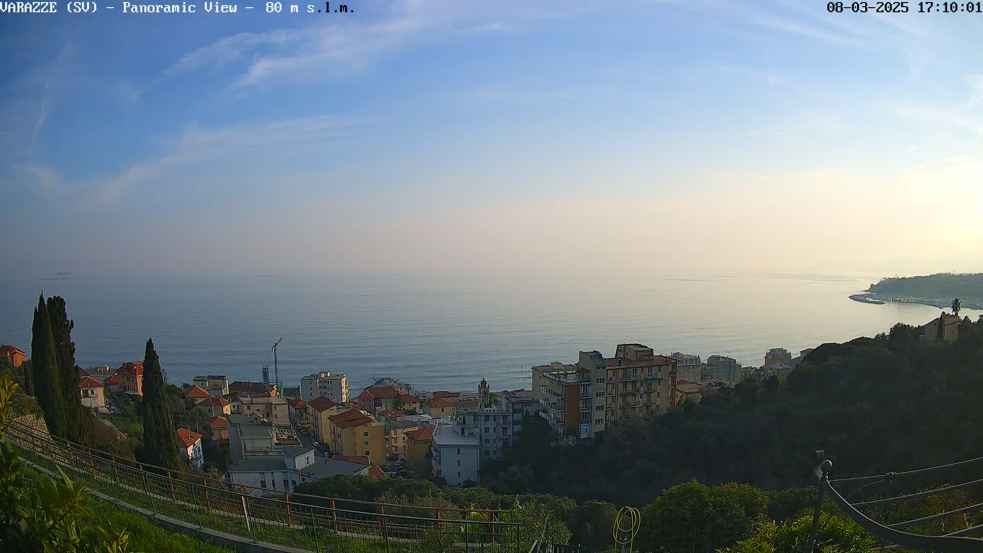 A coastal town with multi-story buildings nestled on a hillside overlooking a calm sea under a clear, light blue sky.