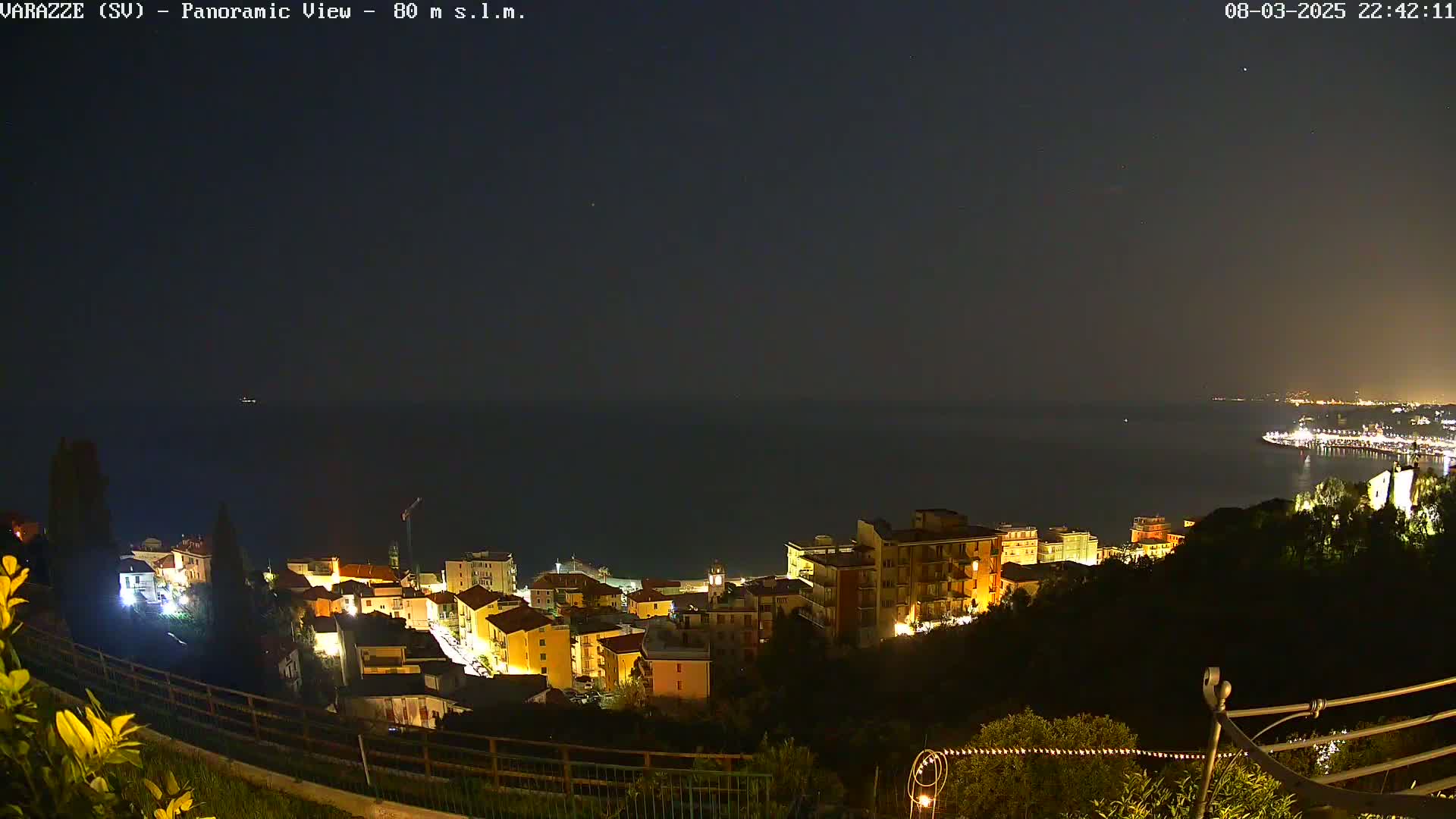 A nighttime panoramic view shows a coastal town with illuminated buildings overlooking a dark sea under a clear sky.