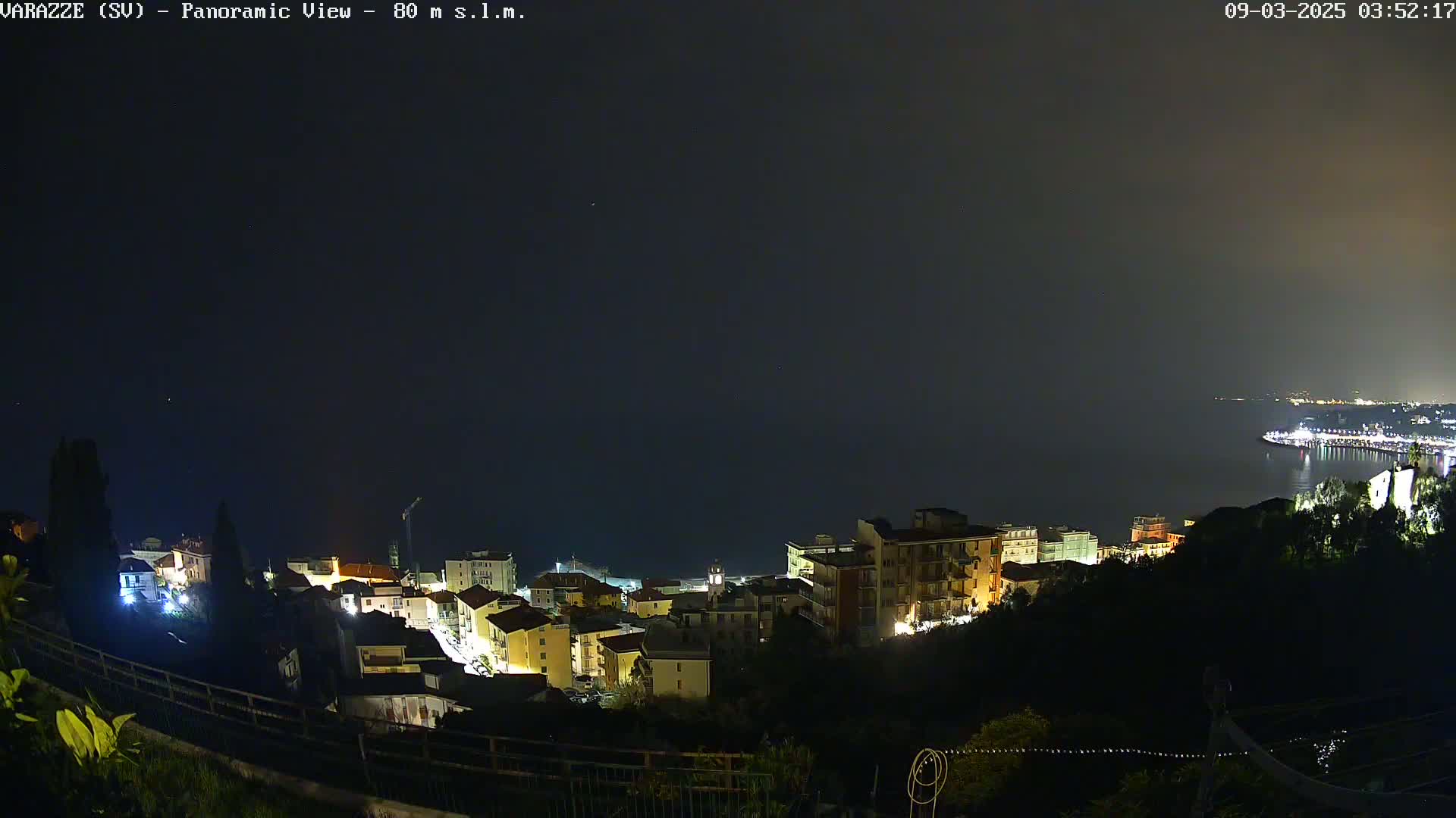 A nighttime panoramic view of a coastal town, illuminated with lights, overlooking a dark body of water under a clear, dark sky.