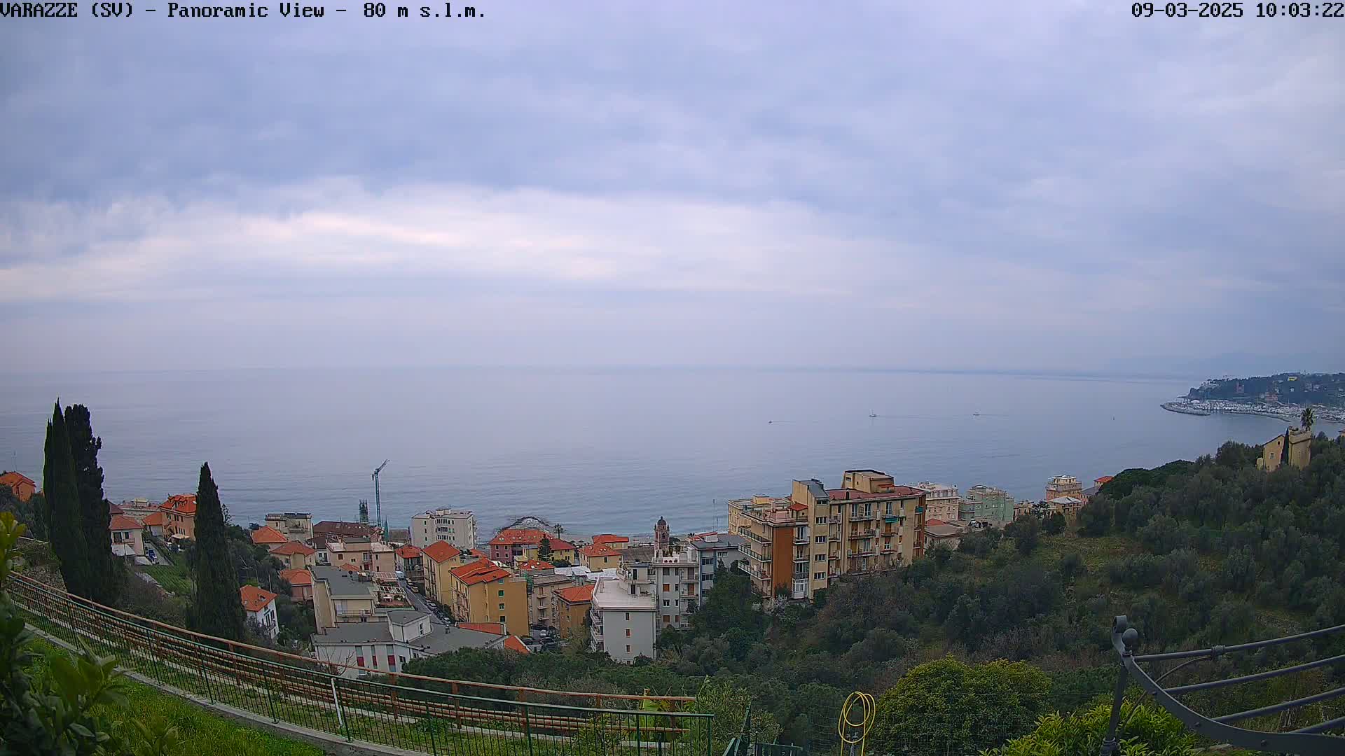 A panoramic view of a coastal town on a cloudy day, showing buildings on a hillside overlooking a calm sea.