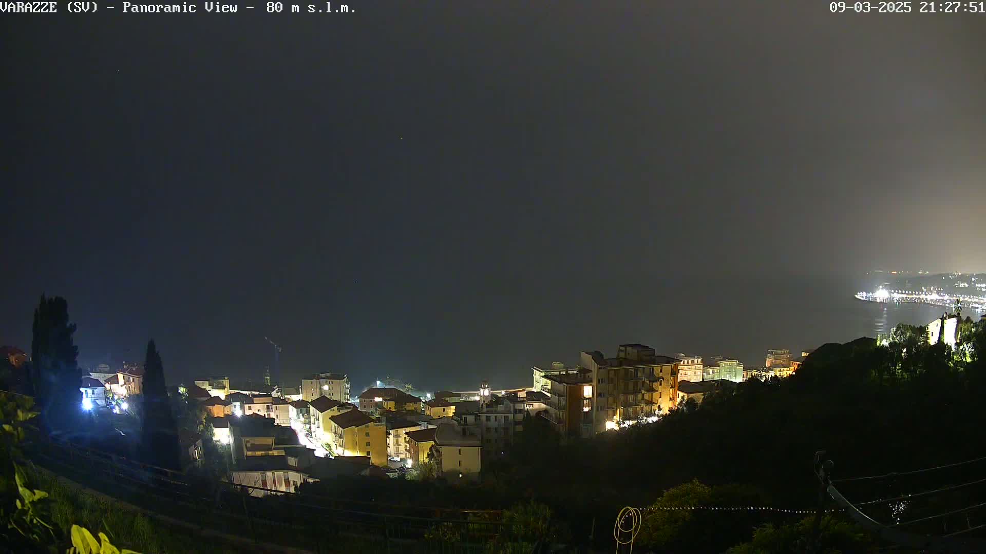 A nighttime panoramic view of a coastal town illuminated by lights, under a dark, hazy sky.