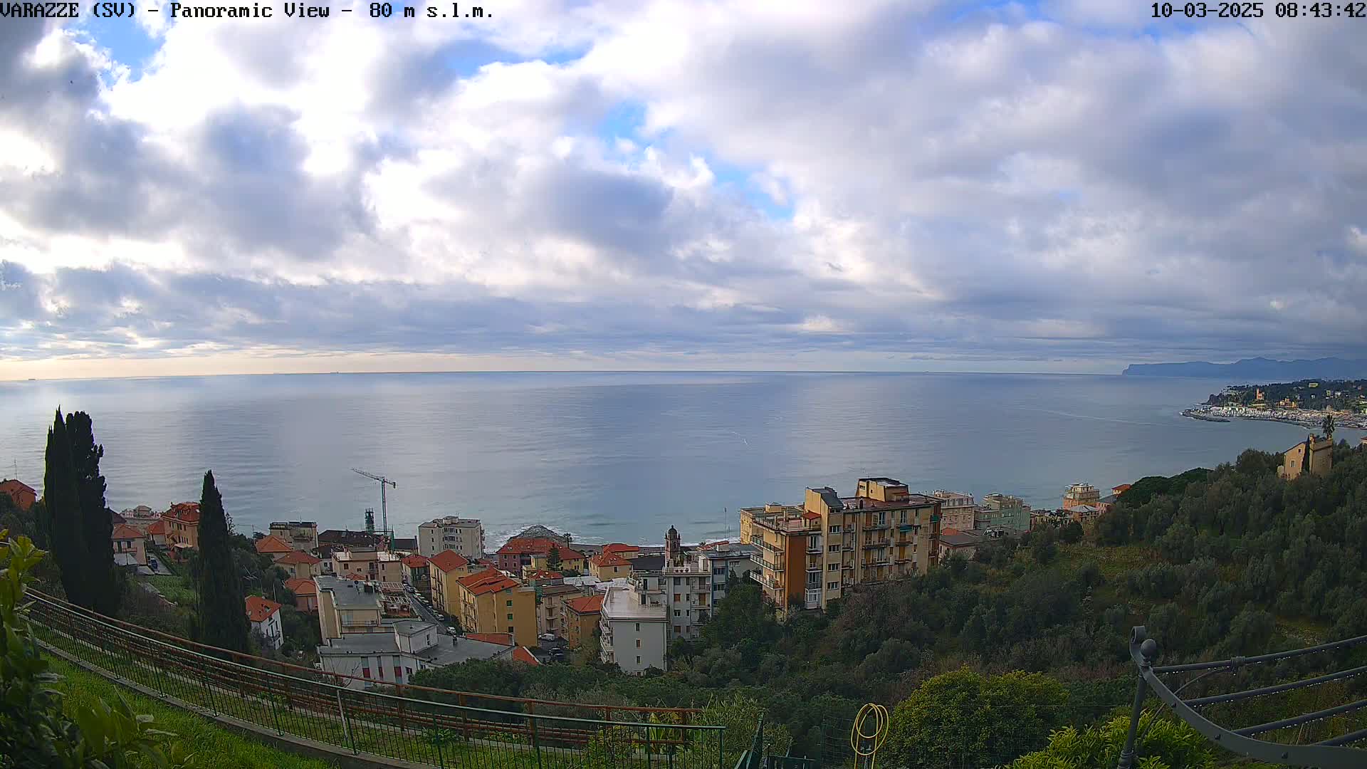 A panoramic view of a coastal town nestled on a hillside overlooking a calm sea under a partly cloudy sky.