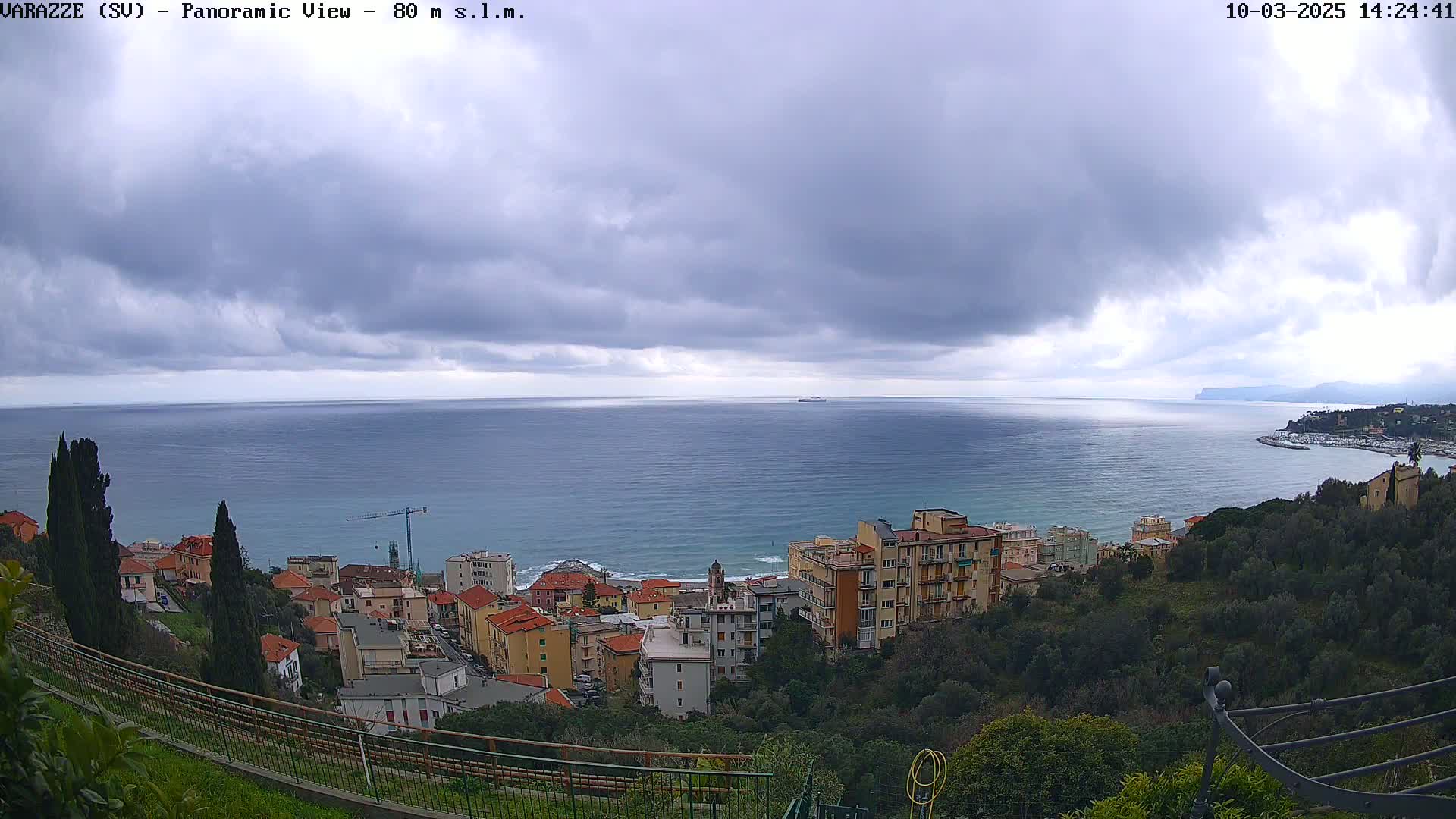 A coastal town with multi-story buildings is nestled on a hillside overlooking a calm sea under a cloudy sky.