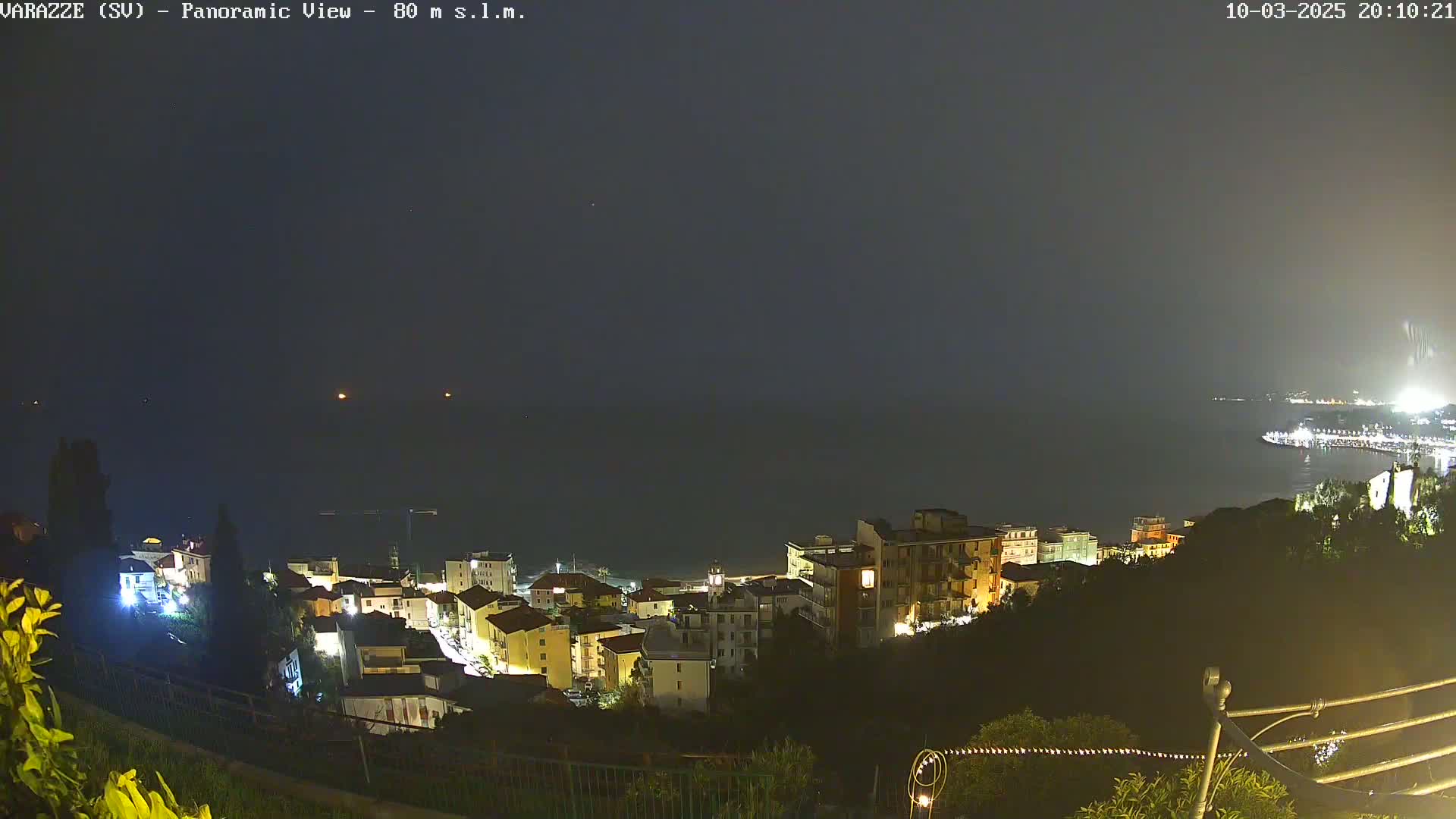 A nighttime, panoramic view of a coastal town illuminated with lights, overlooking a dark body of water under a hazy sky.