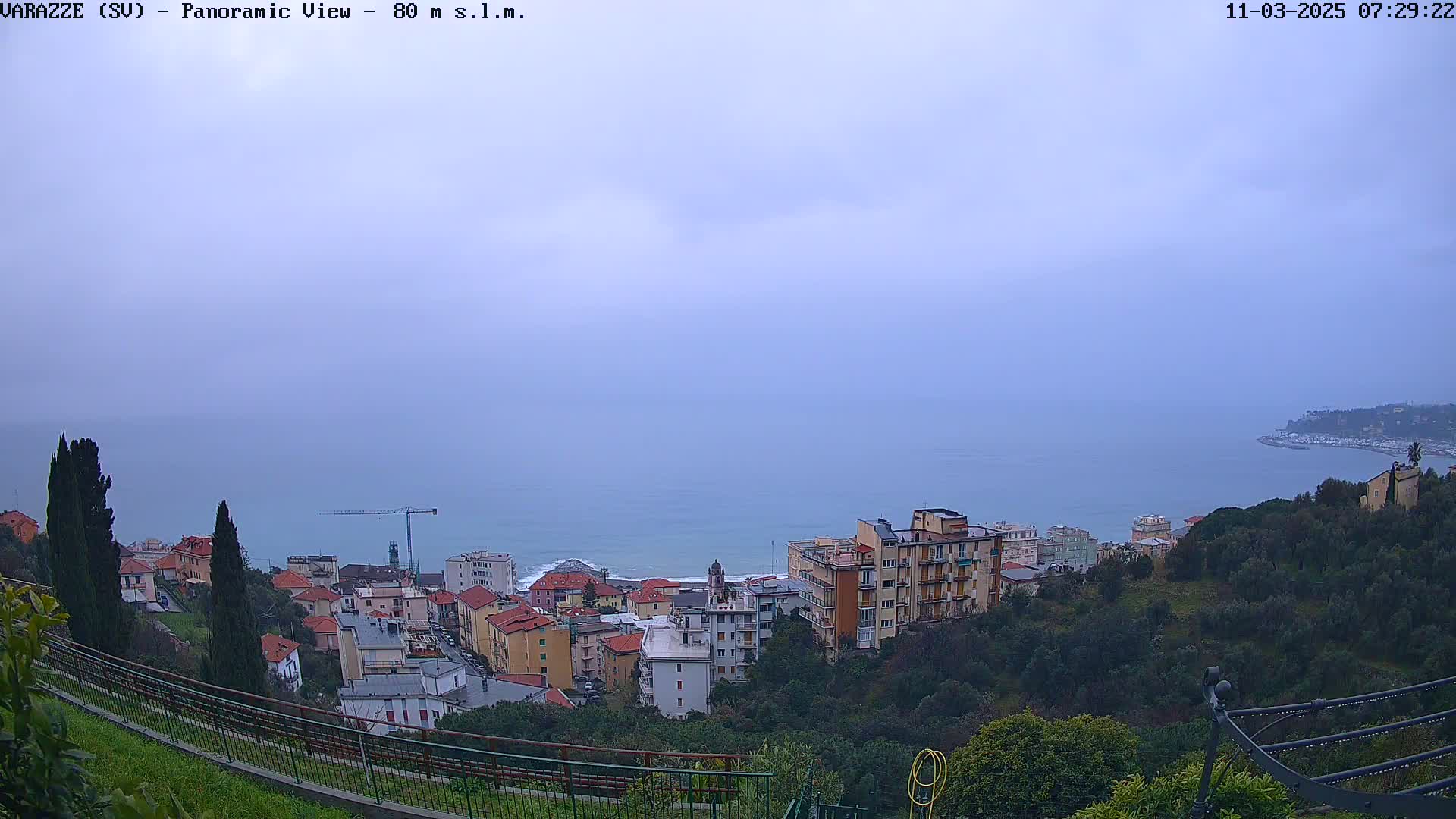 A coastal town nestled on a hillside overlooks a calm sea under an overcast sky.