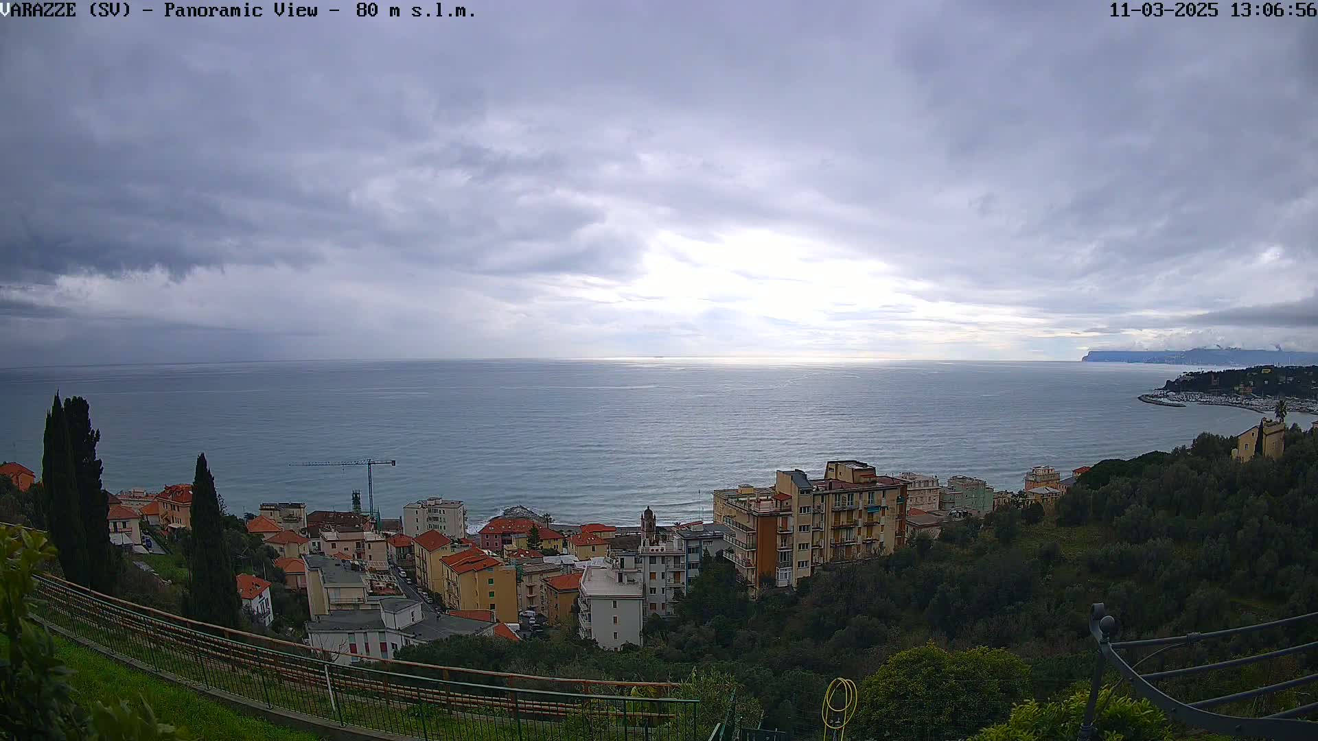 A coastal town with multi-story buildings is seen from an elevated vantage point overlooking a calm sea under a mostly cloudy sky.
