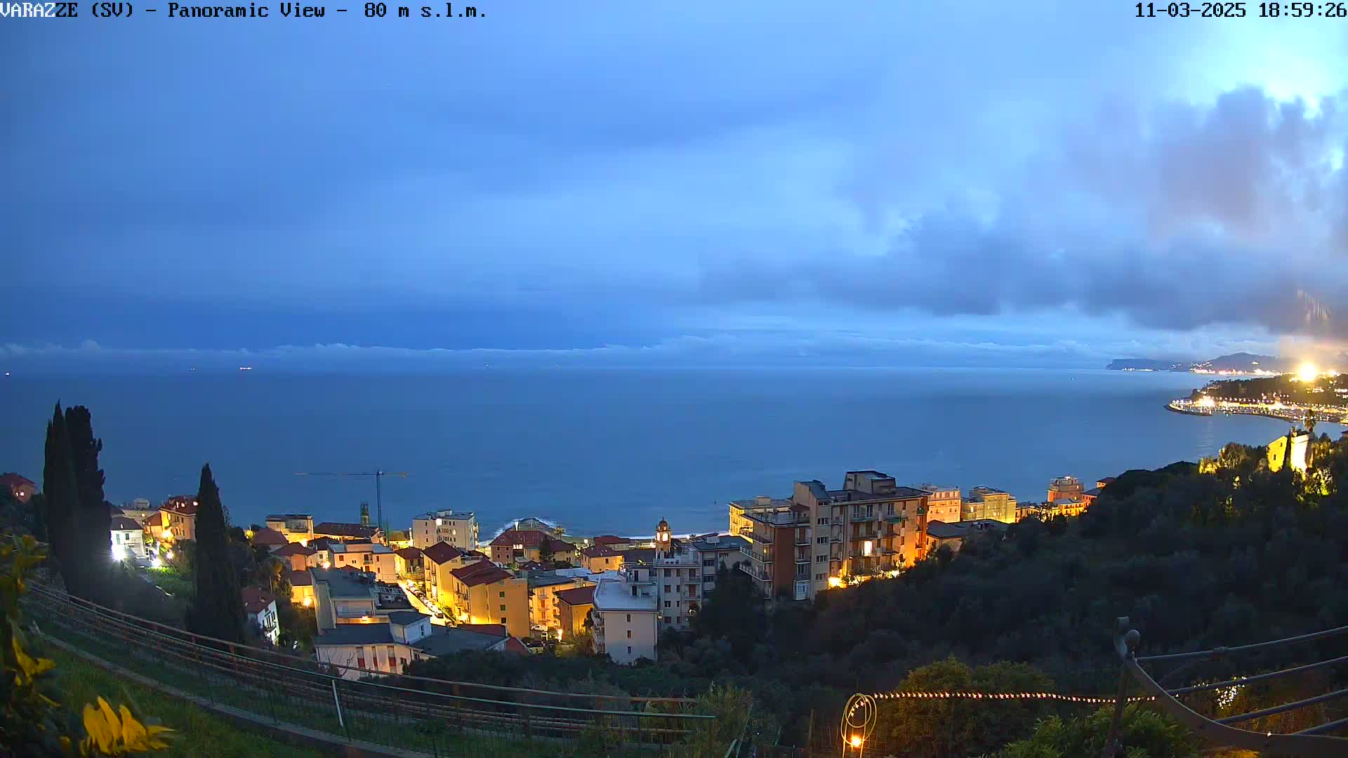 A coastal town with lit buildings sits on a hillside overlooking a calm, dark-blue sea under a mostly cloudy twilight sky.
