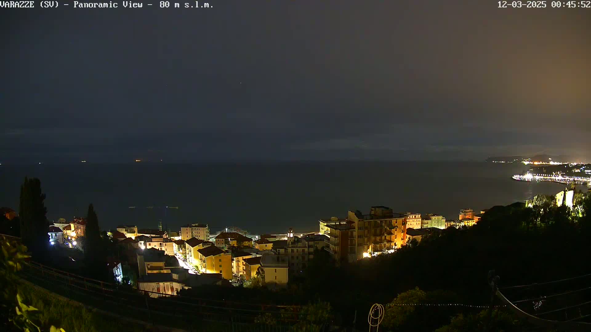 A nighttime panoramic view shows a coastal town with illuminated buildings overlooking a dark sea under a cloudy sky.