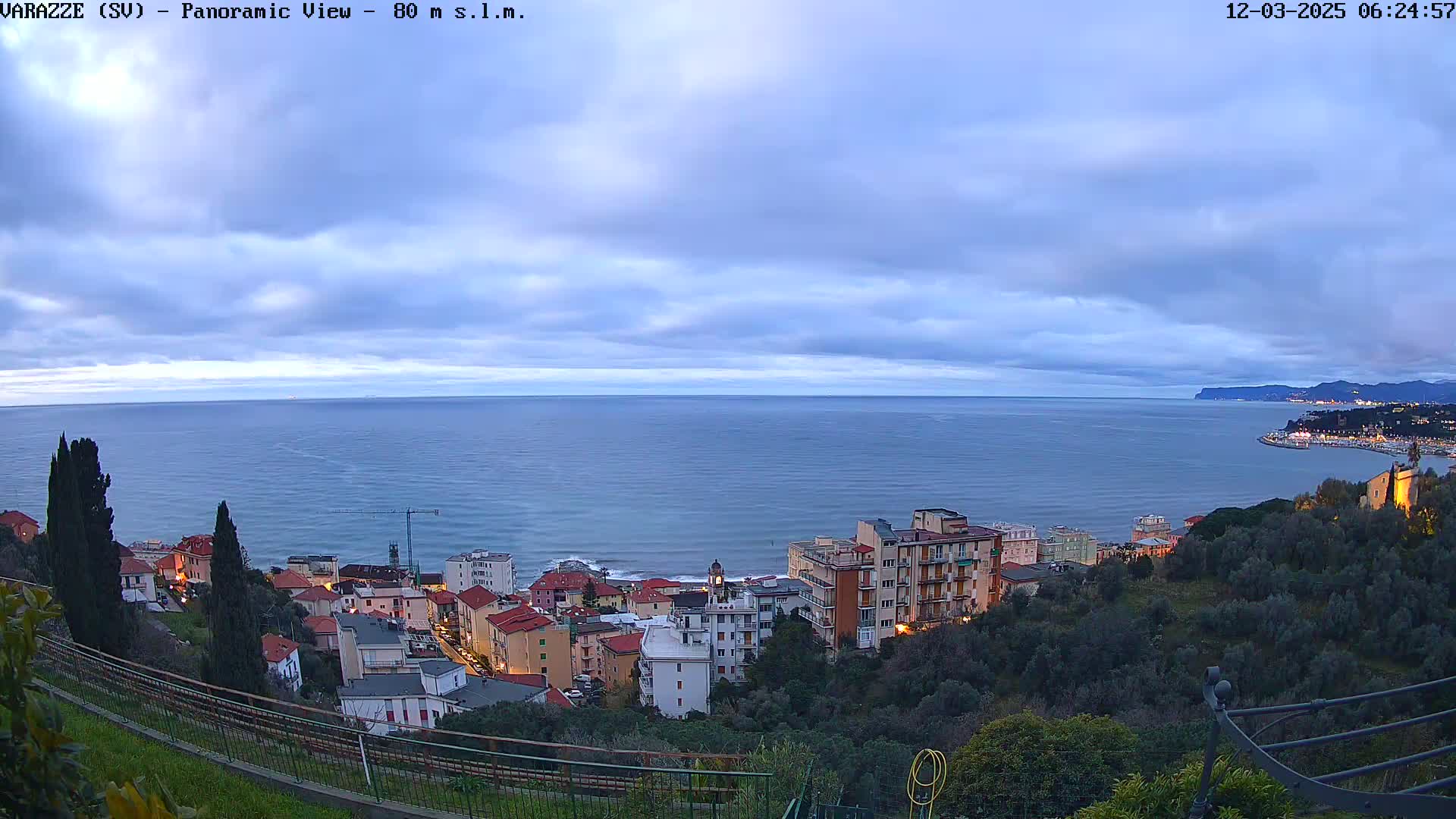 A coastal town with multi-story buildings is nestled on a hillside overlooking a calm sea under a mostly cloudy, twilight sky.