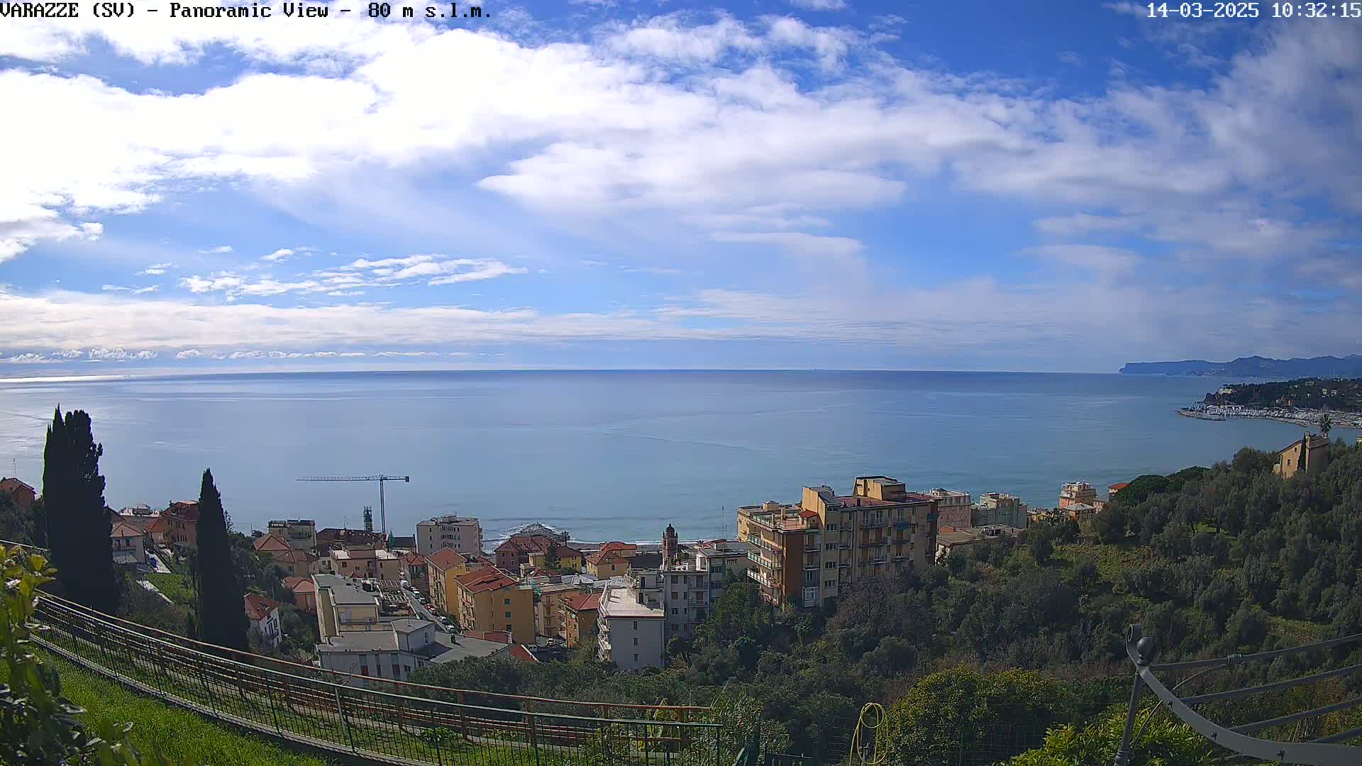 A partly cloudy day reveals a coastal town nestled on a hillside overlooking a calm sea, with a construction crane visible near the shoreline.