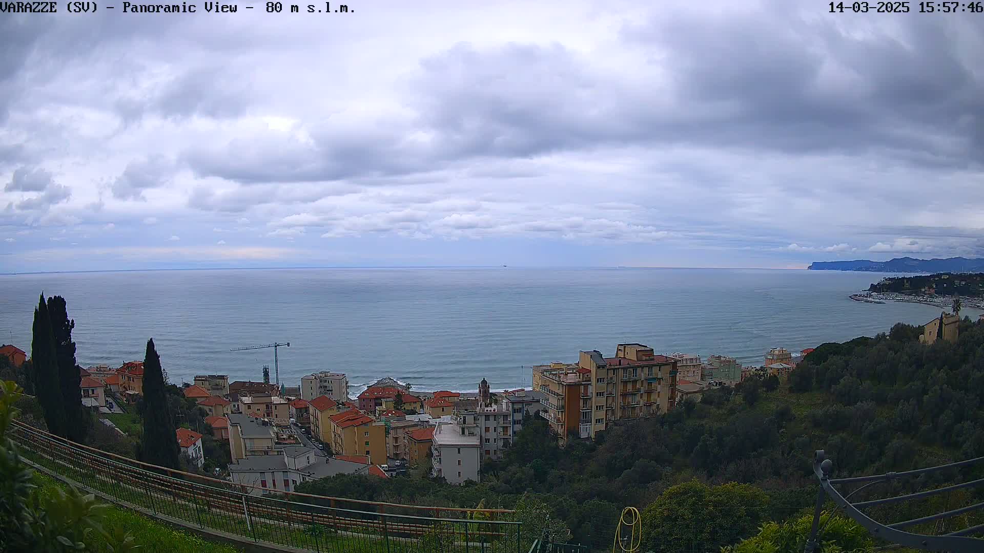 A panoramic view of a coastal town under a cloudy sky shows a calm sea extending to the horizon, with buildings nestled on a hillside overlooking the water.