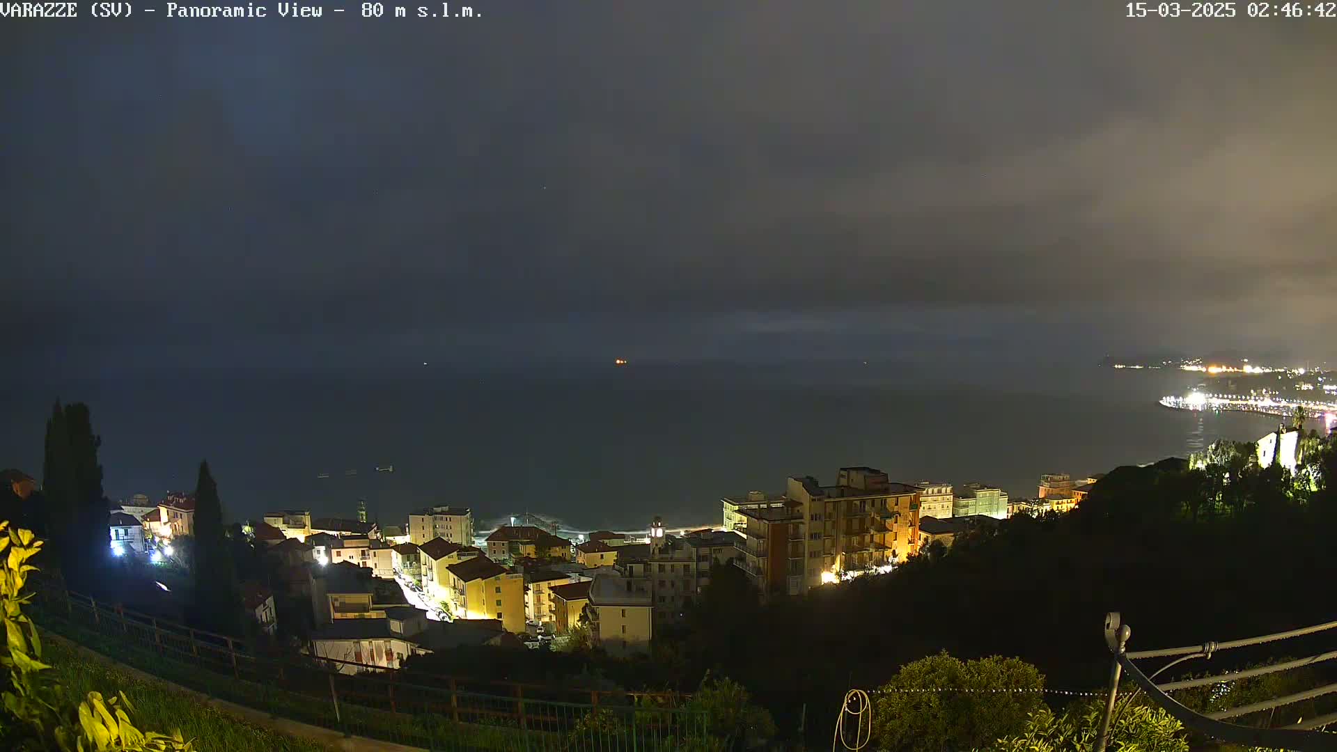 A nighttime panoramic view of a coastal town with its lights illuminated under a cloudy sky, overlooking a dark sea.