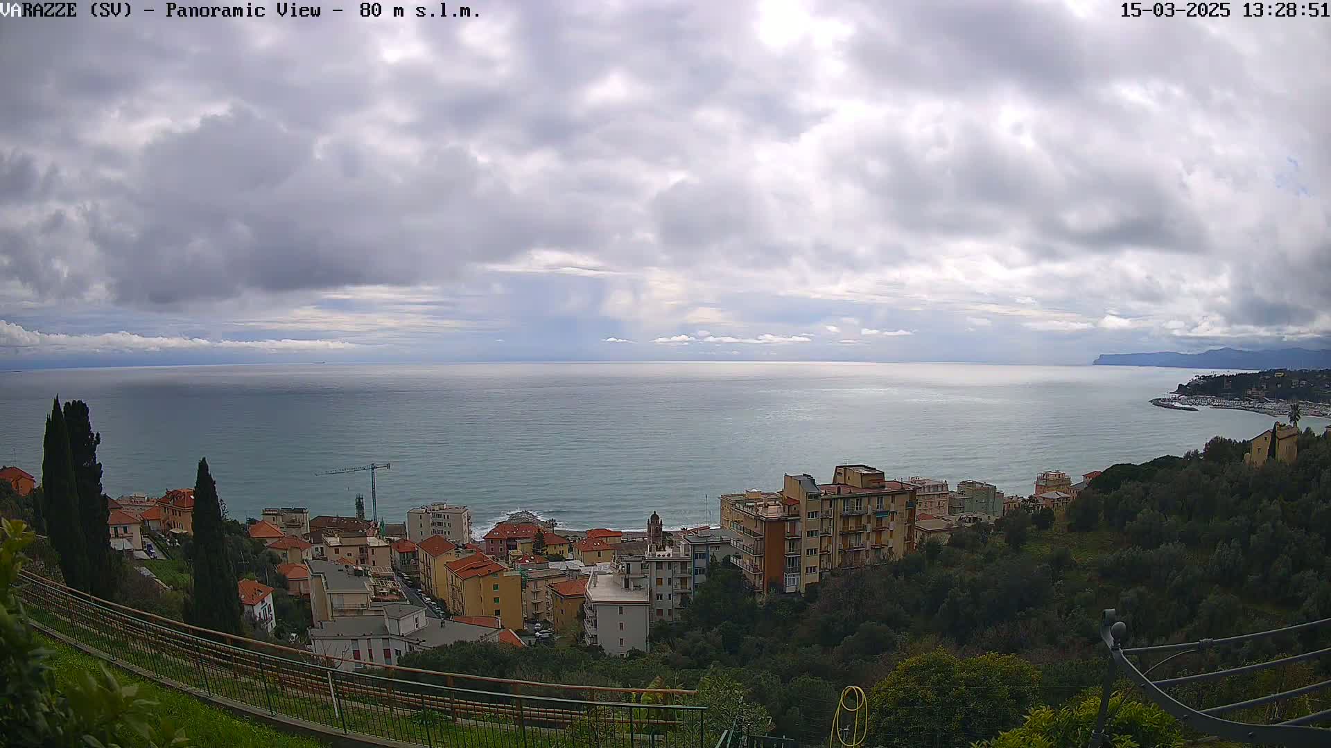 A coastal town with multi-story buildings is nestled on a hillside overlooking a calm sea under a cloudy sky.