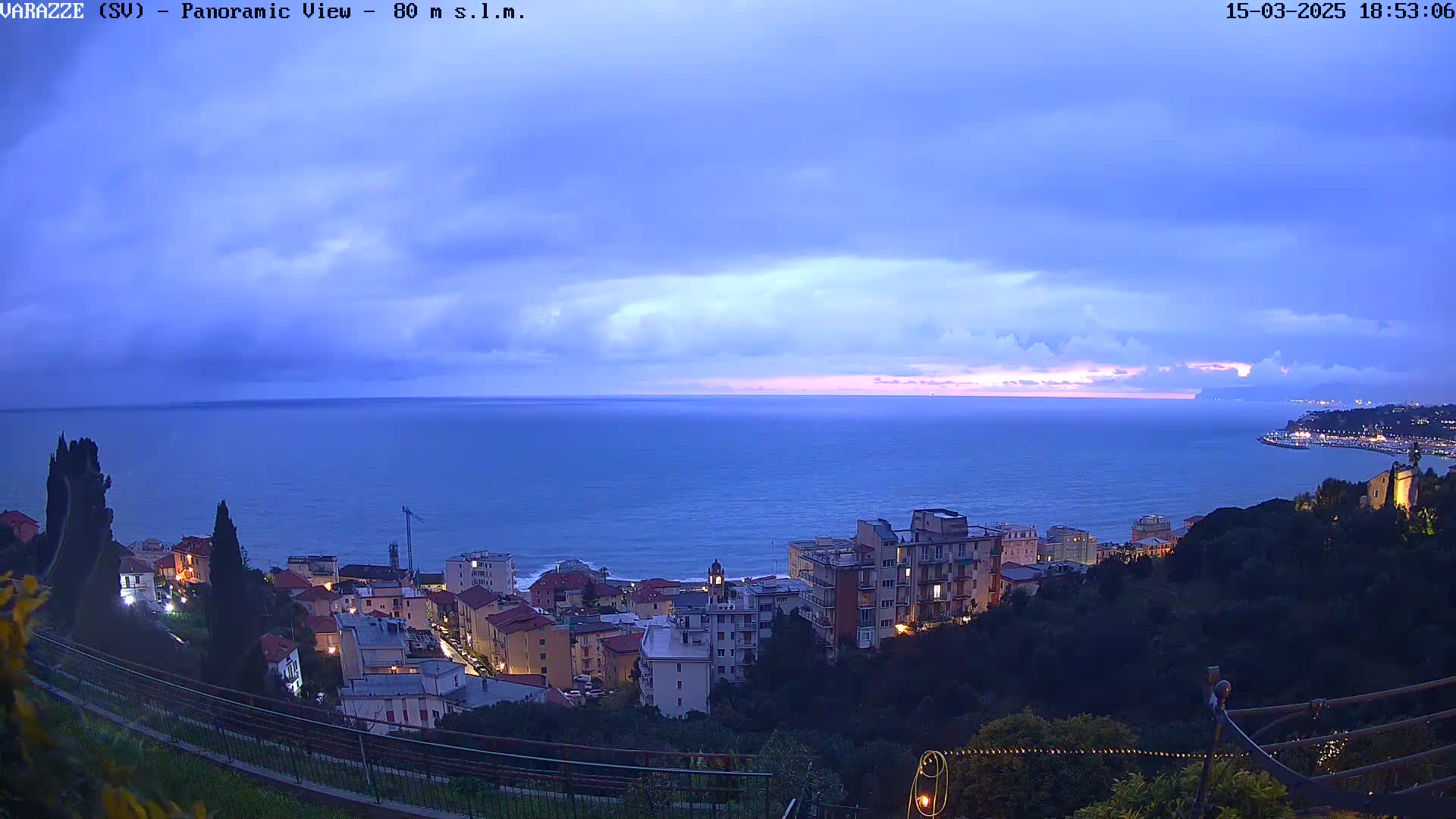 A panoramic view of a coastal town at dusk under a cloudy sky shows the town's lights illuminated against the dark blue sea.