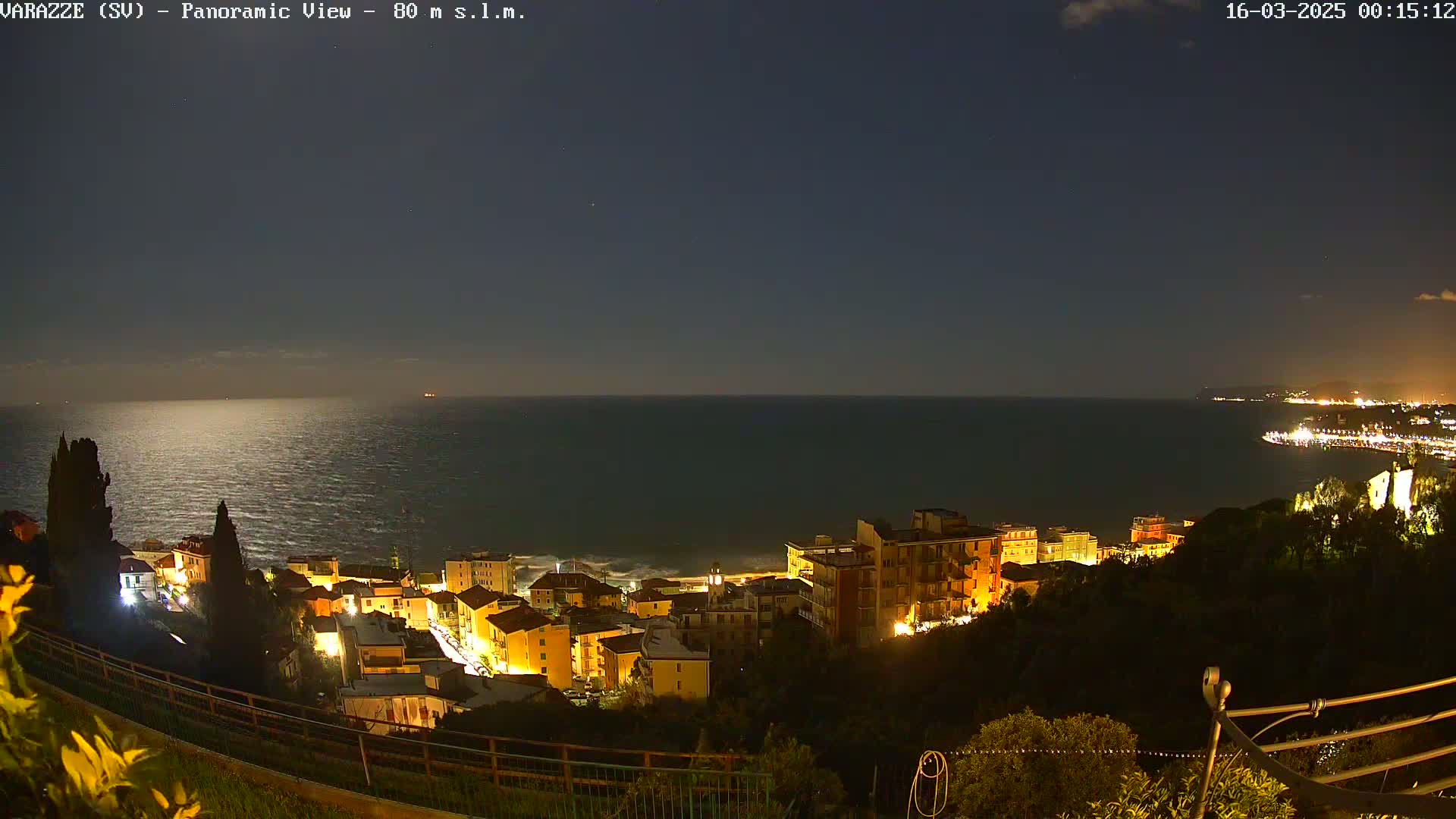 A nighttime panoramic view shows a coastal town illuminated, situated on a hillside overlooking a dark, calm sea under a mostly clear sky.