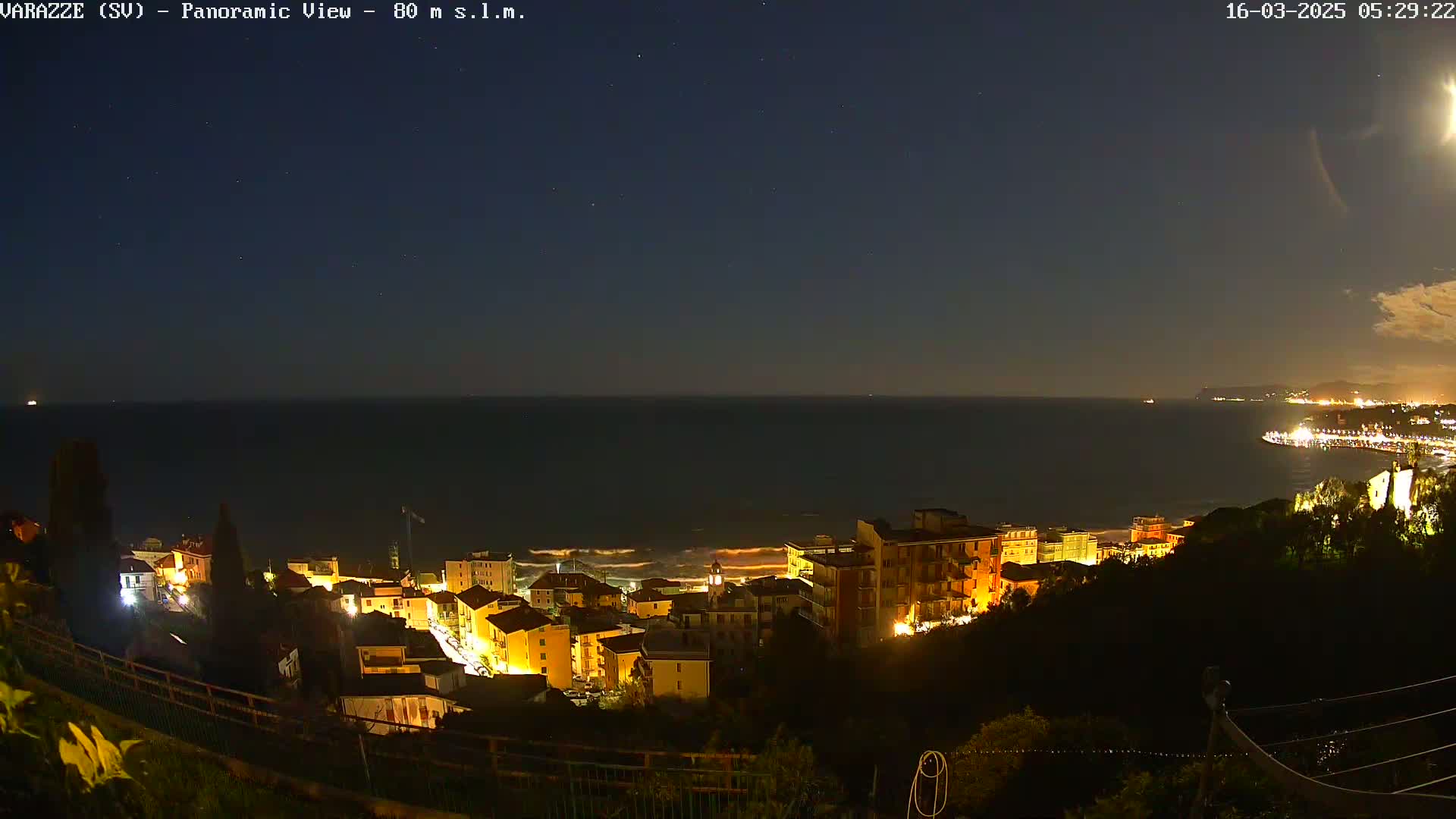 A nighttime panoramic view shows a coastal town illuminated with lights, overlooking a dark sea under a clear, starry sky.