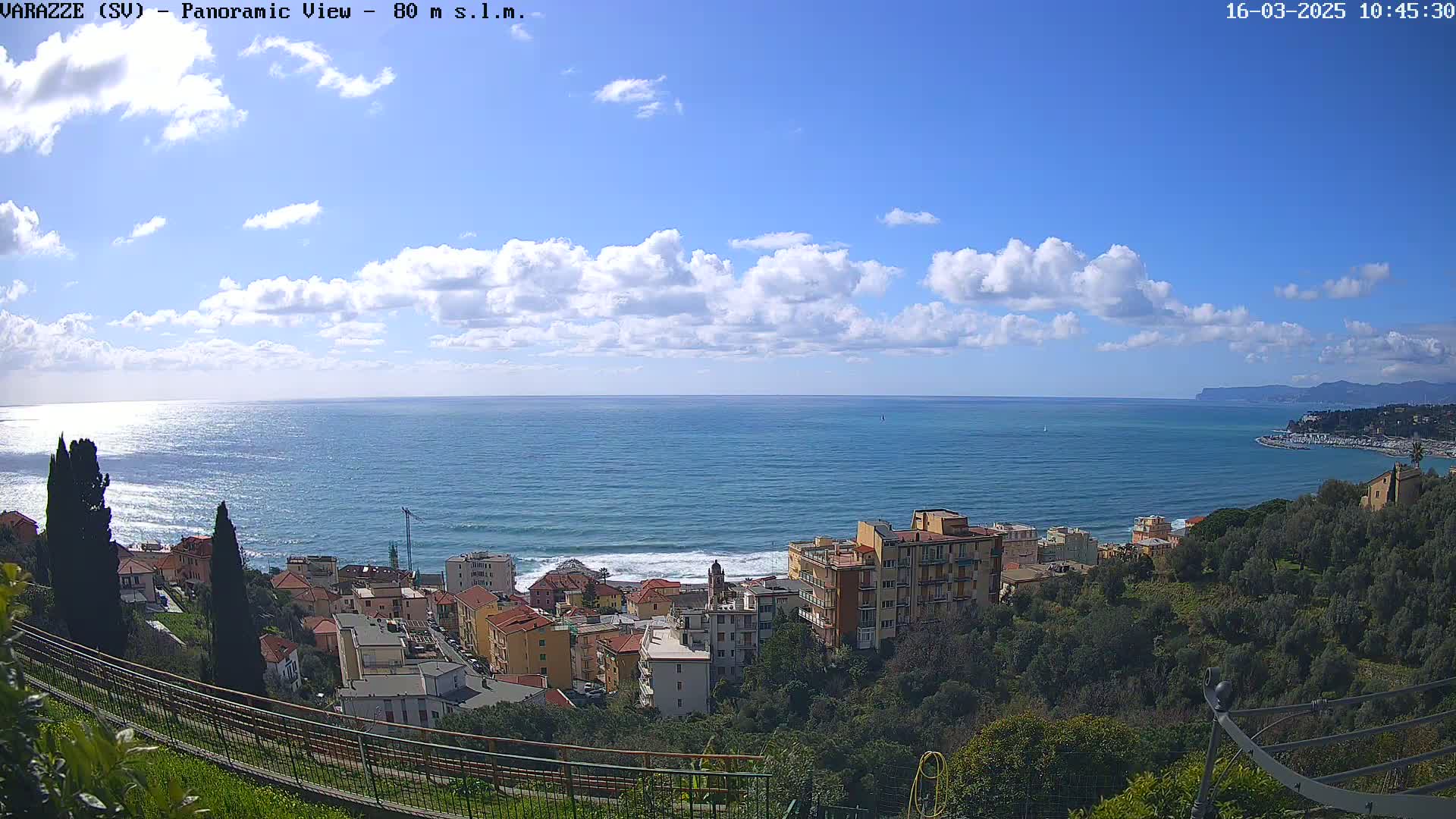A panoramic view of a coastal town under a partly cloudy sky shows a calm sea with small waves breaking on the shore.
