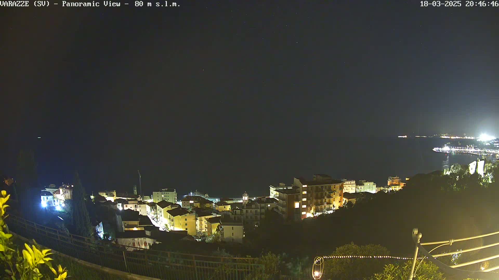 A nighttime, panoramic view shows a coastal town illuminated, with a dark sea extending to a dimly lit coastline in the distance under a clear, starry sky.
