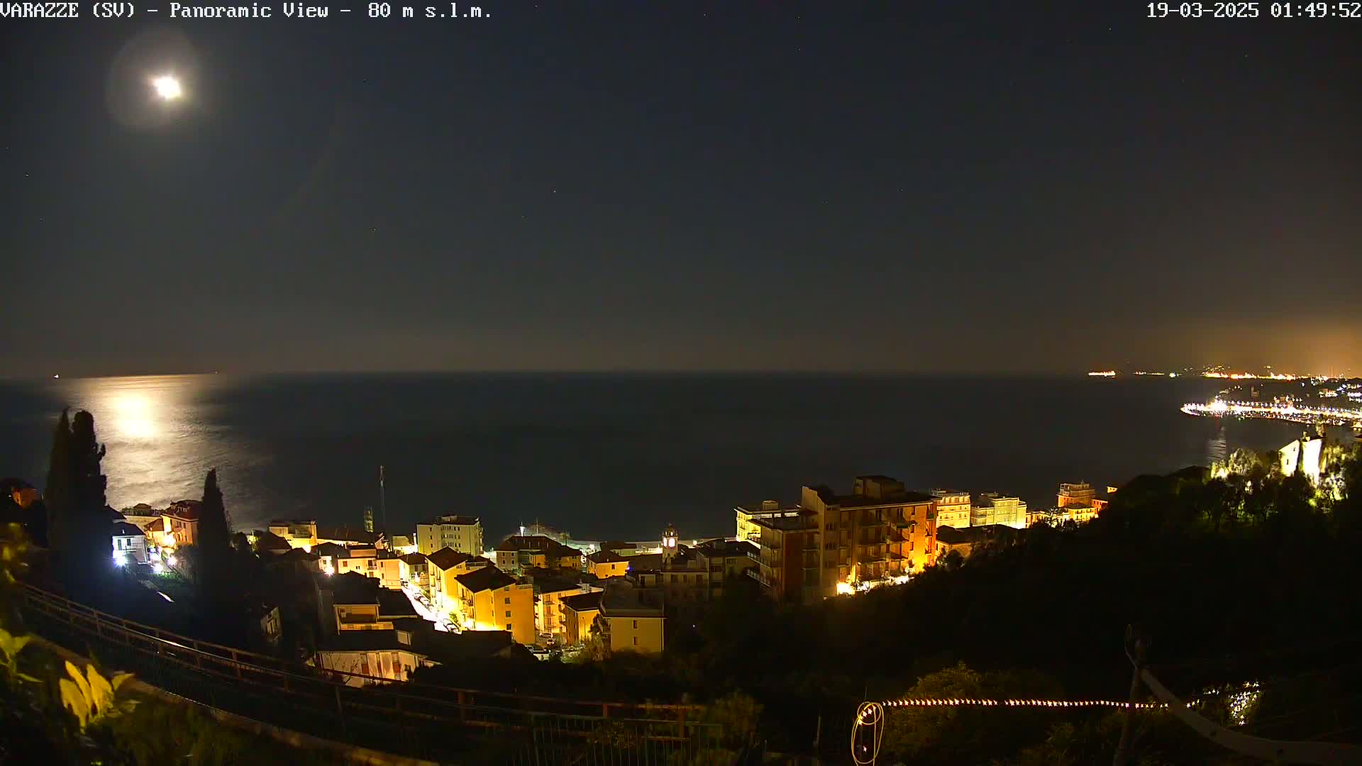 A moonlit nighttime view of a coastal town with buildings illuminated, overlooking a calm sea under a clear sky.