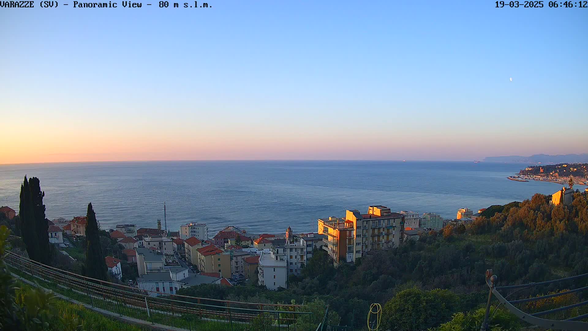 A coastal town with multi-story buildings sits on a hillside overlooking a calm ocean at sunset under a clear sky.