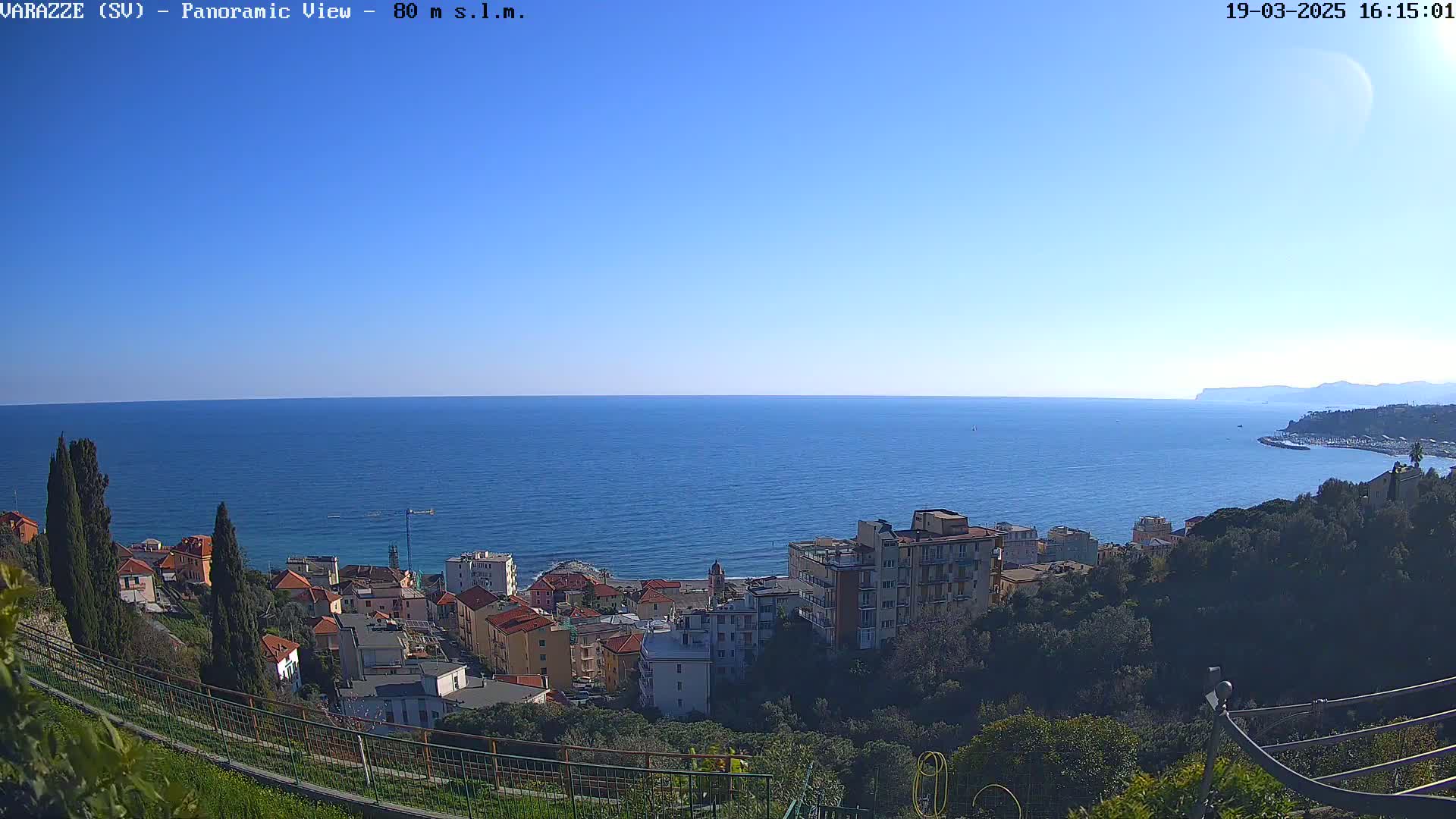 A panoramic view shows a coastal town nestled on a hillside overlooking a calm, blue sea under a clear, sunny sky.