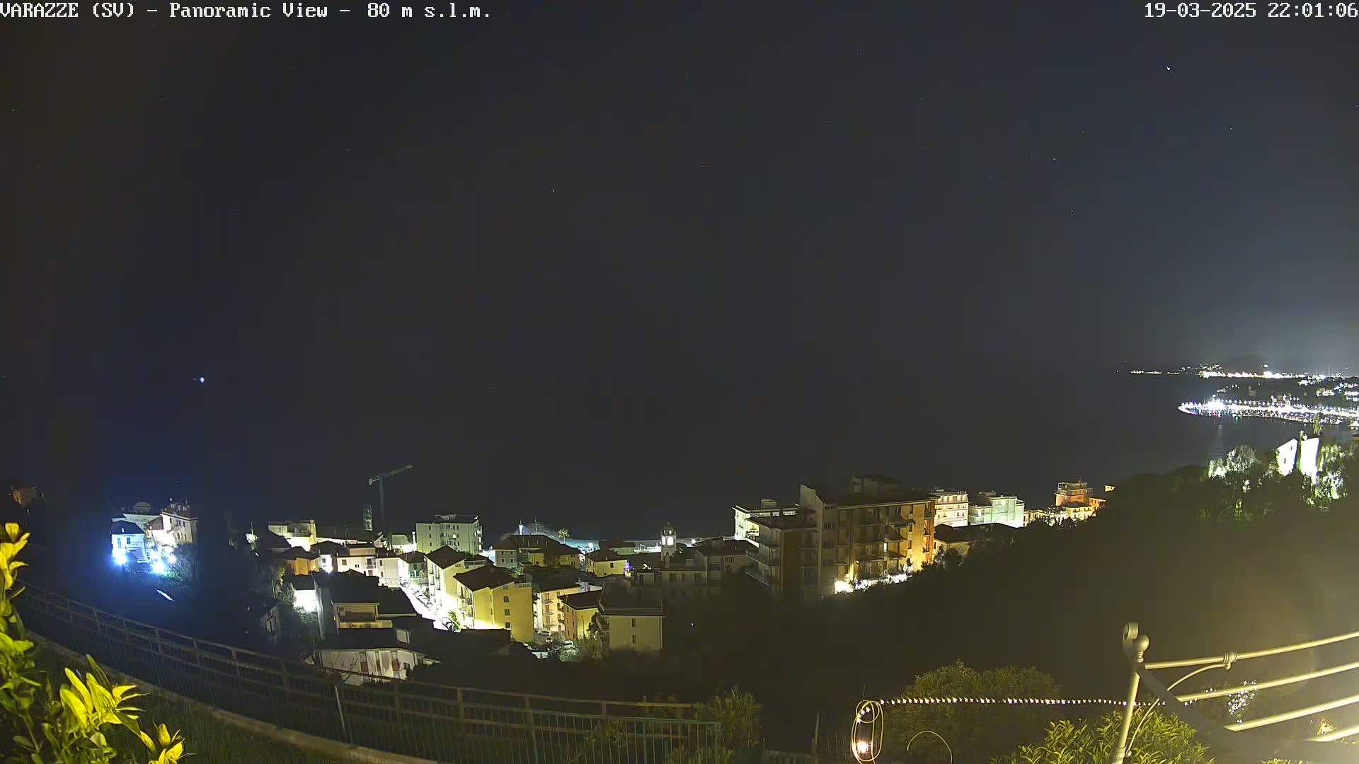 A nighttime panoramic view of a coastal town illuminated with lights, under a clear, starry sky.